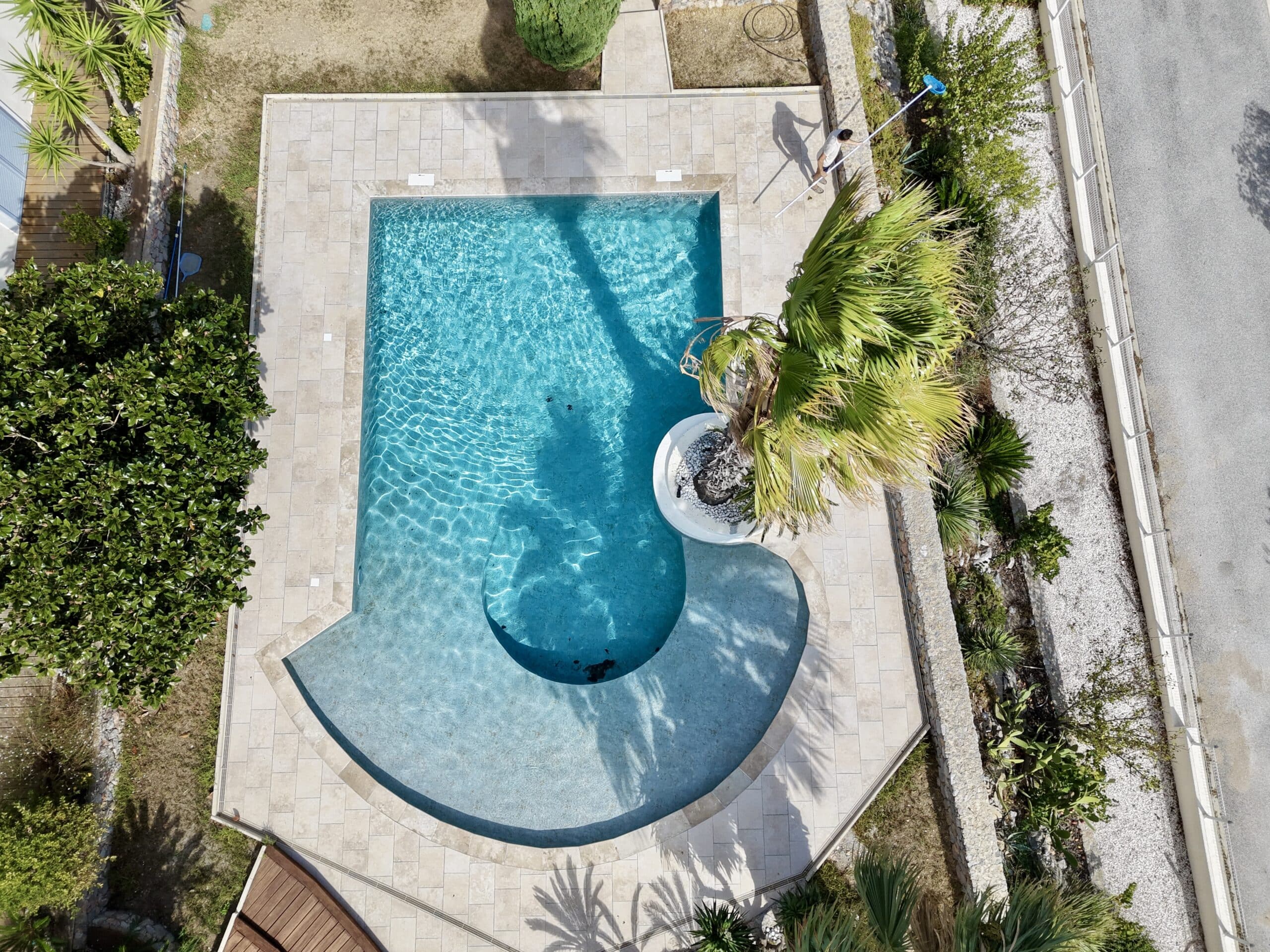 Vue aérienne d'une piscine design bleu clair avec palmier central et personne effectuant le nettoyage.