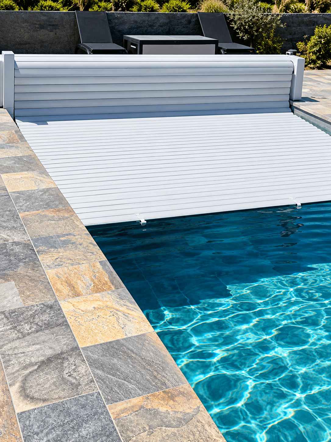 Volet roulant automatique blanc se déployant sur une piscine bleue moderne bordée de dalles en pierre naturelle.