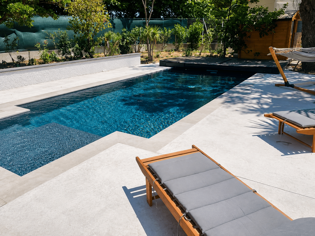 Piscine Moderne avec Terrasse en Pierre et Chaises Longues Piscine moderne aux carreaux bleus, terrasse en pierre blanche, chaises longues en bois et jardin verdoyant.