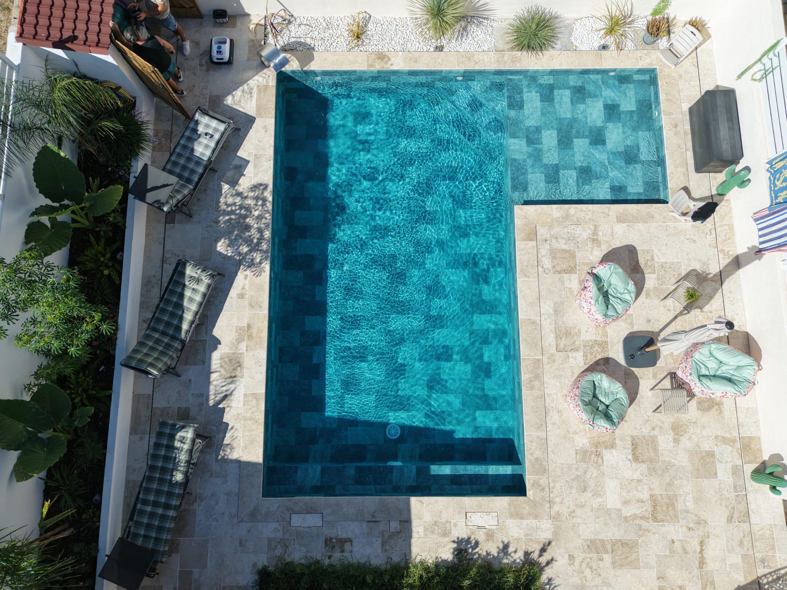 Vue aérienne d'une piscine turquoise entourée d'une terrasse en travertin avec transats et poufs colorés en plein soleil.