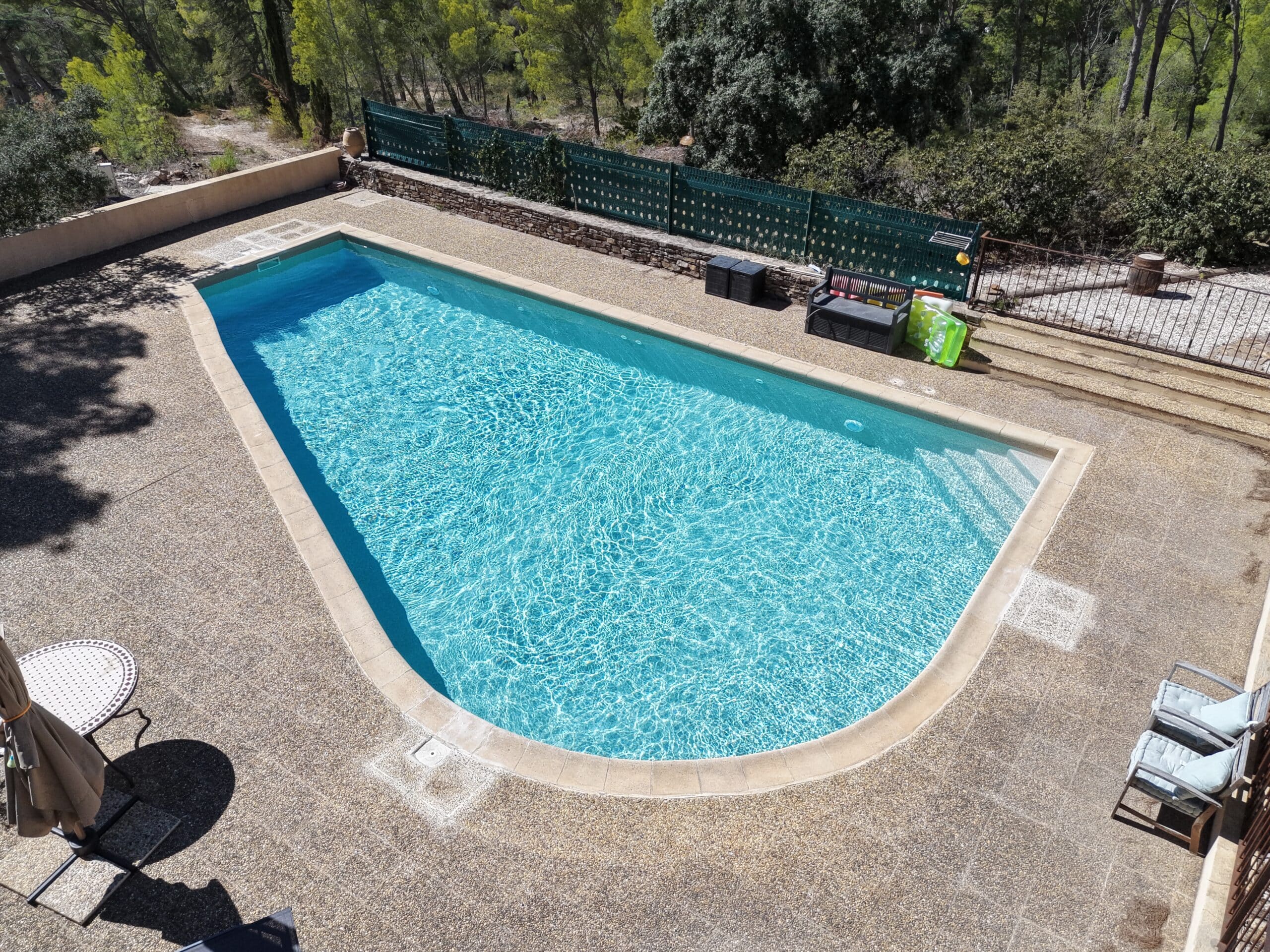 Piscine ensoleillée à l'eau turquoise sur terrasse en pierre et jardin arboré.