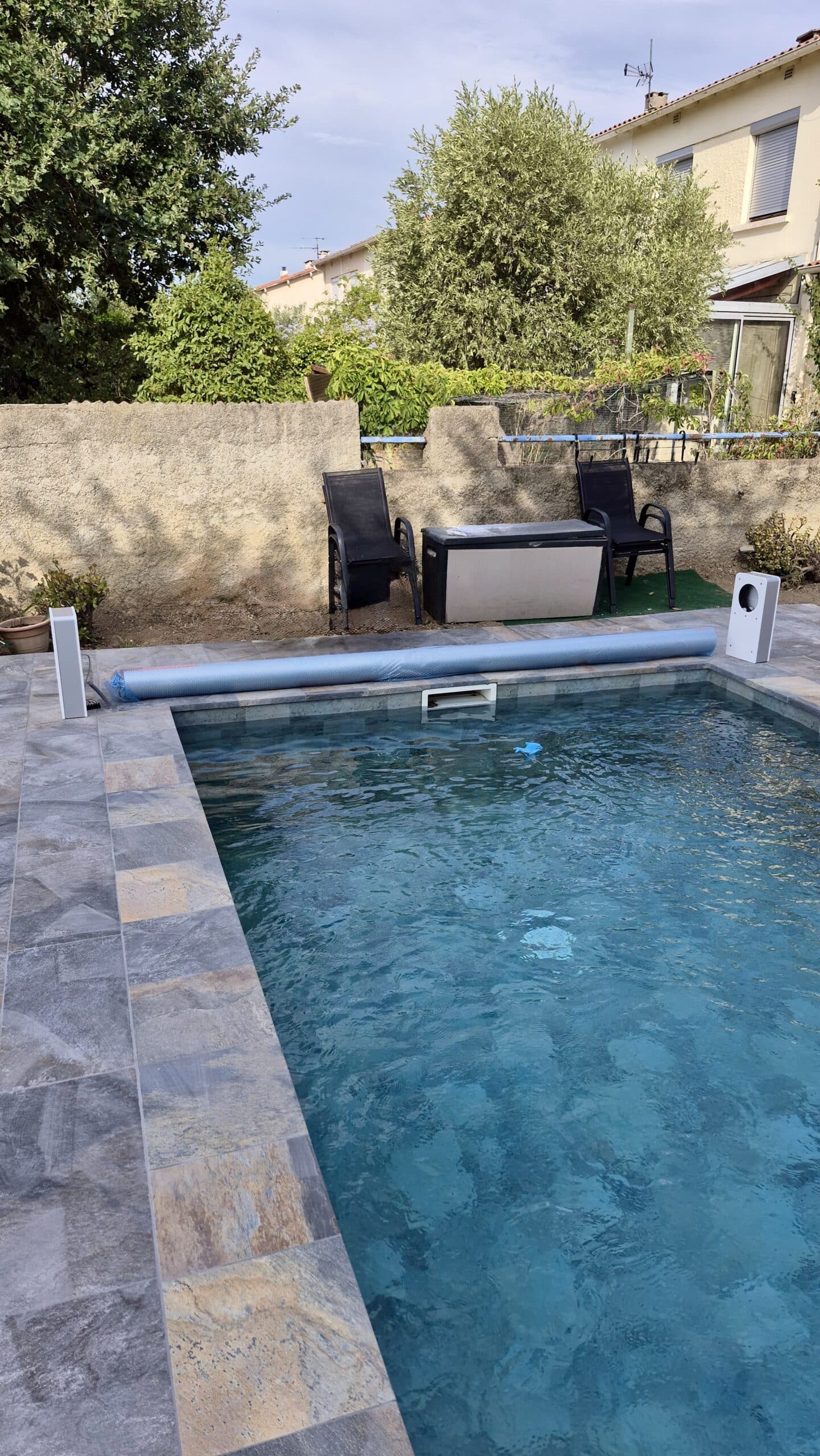 Piscine avec carrelage gris, bâche à bulles enroulée, chaises de jardin noires et végétation en arrière-plan.