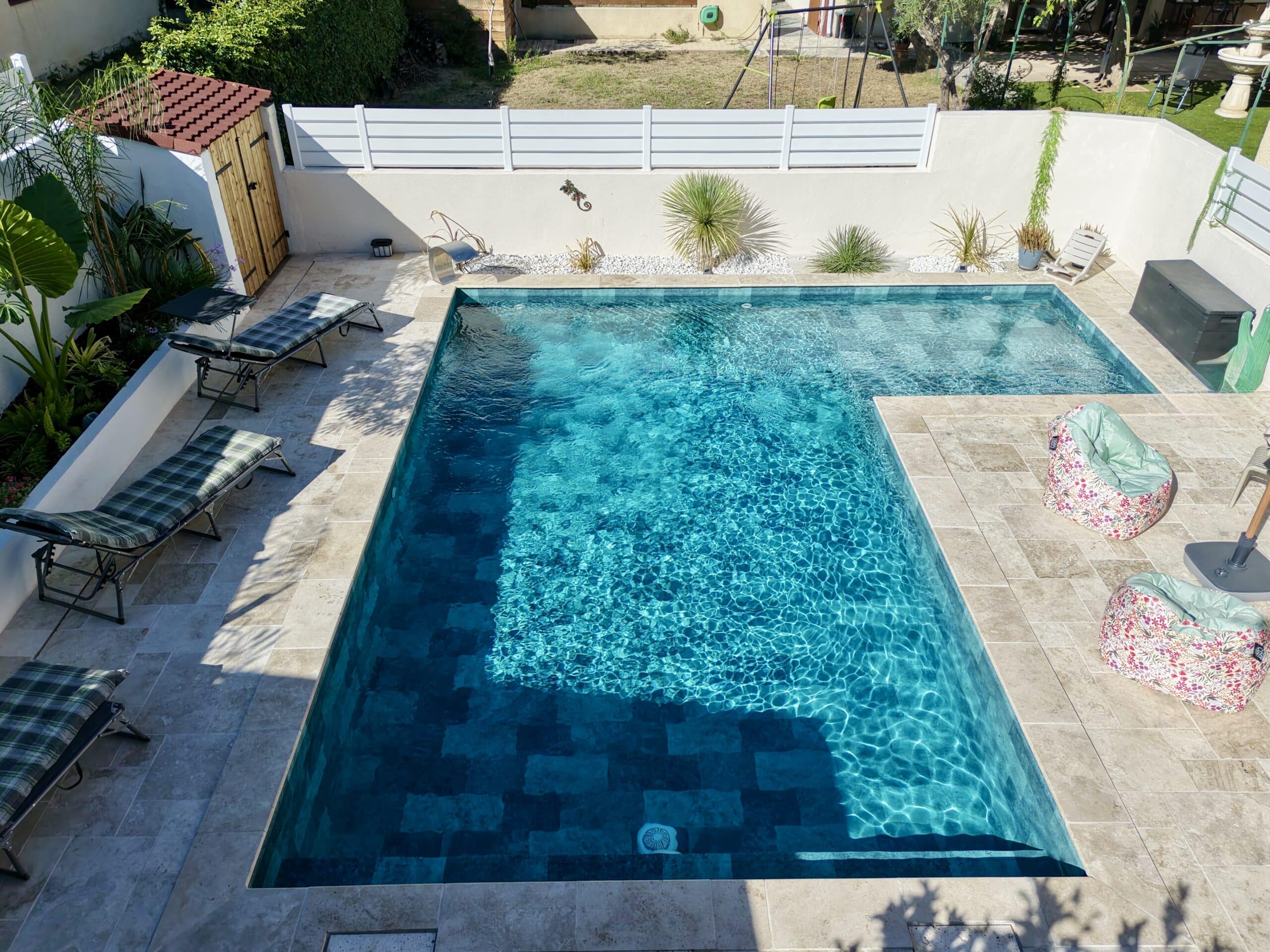 Piscine rectangulaire moderne avec eau bleue cristalline, terrasse en travertin, transats et poufs dans un jardin ensoleillé.
