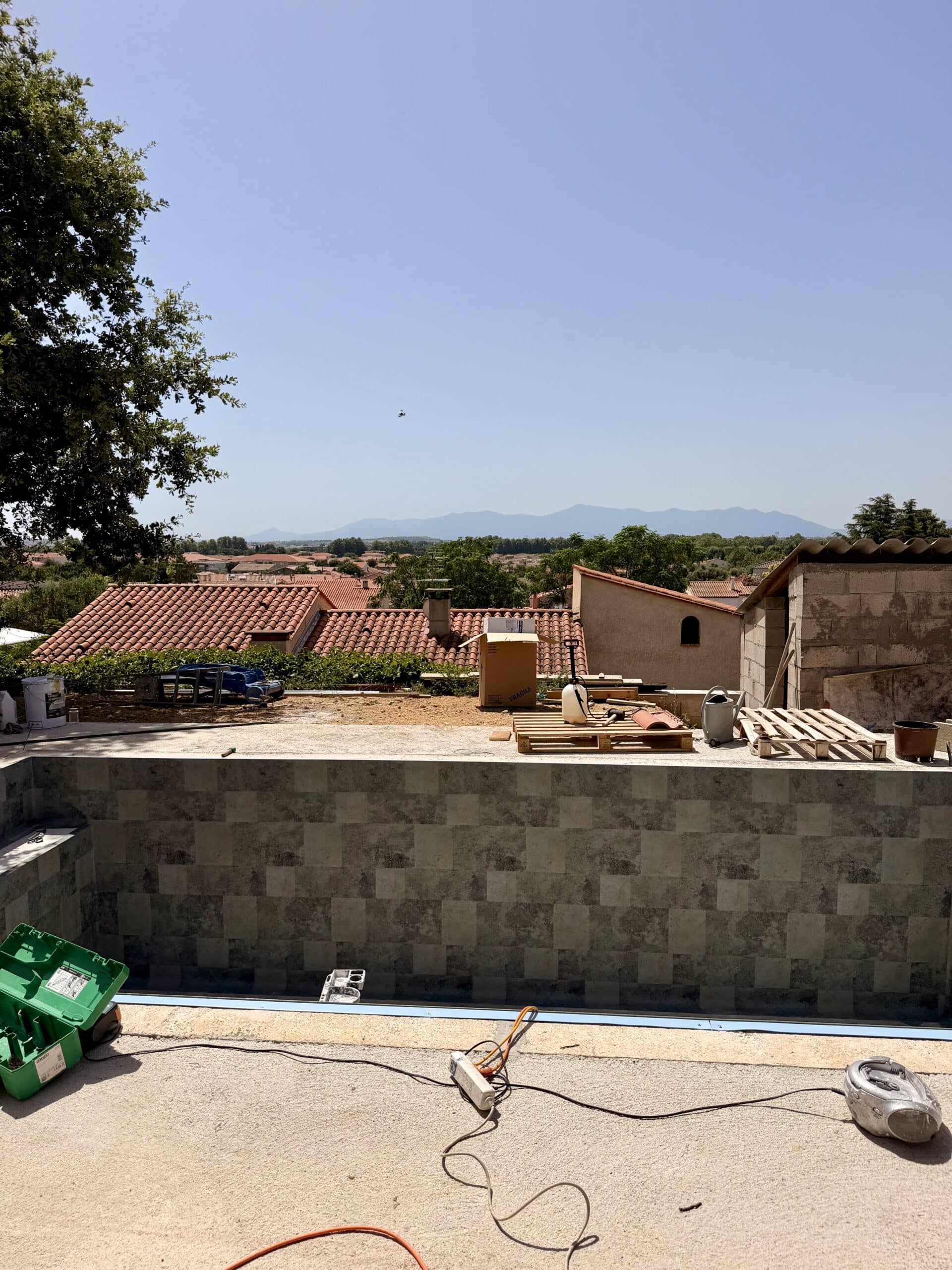 Piscine en cours de carrelage avec vue panoramique sur les toits et montagnes, chantier marqué fragile.