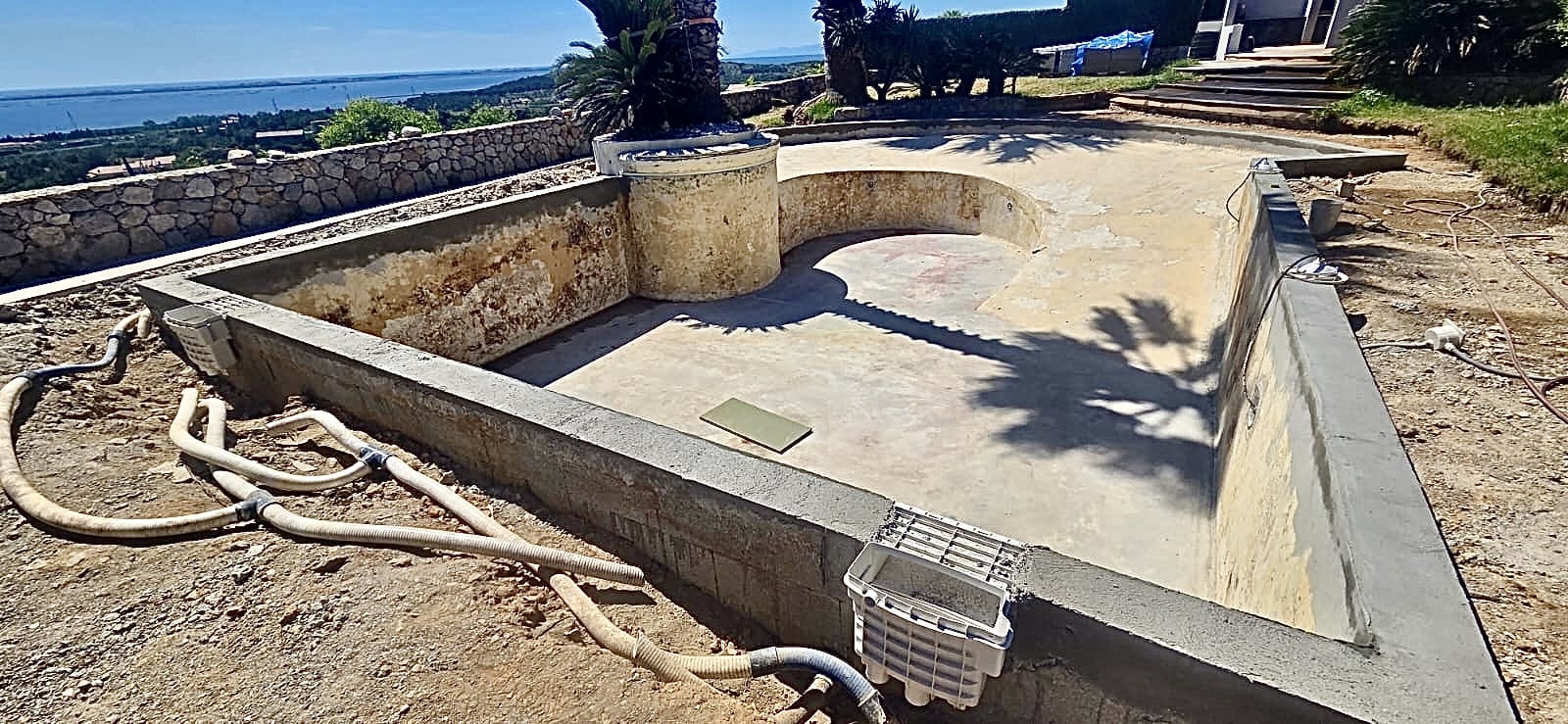 Chantier de rénovation d'une piscine en béton avec vue sur mer, tuyaux de filtration et skimmers apparents.