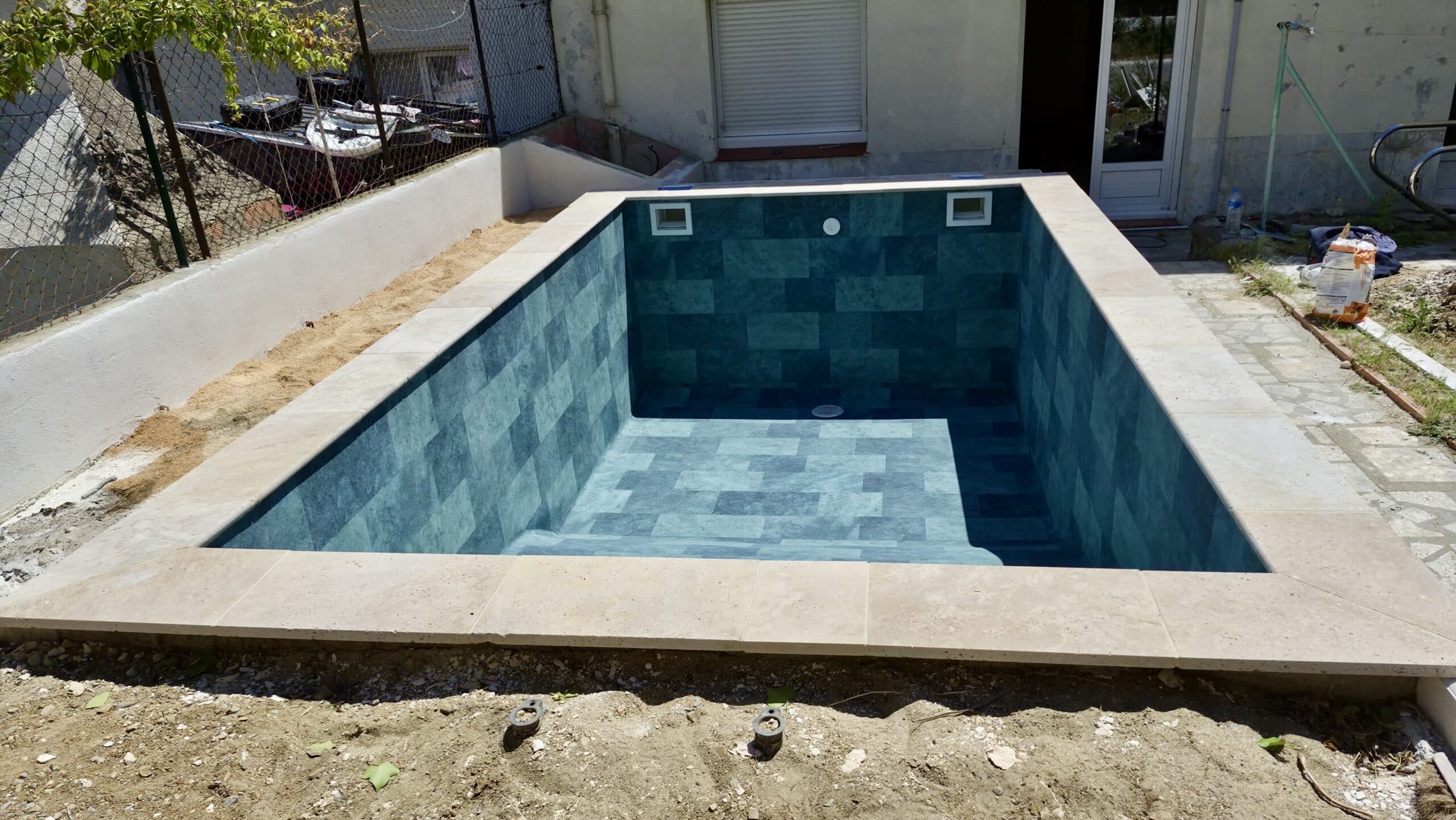 Petite piscine enterrée aux carreaux bleu canard et margelles claires en pierre, chantier de rénovation de jardin.