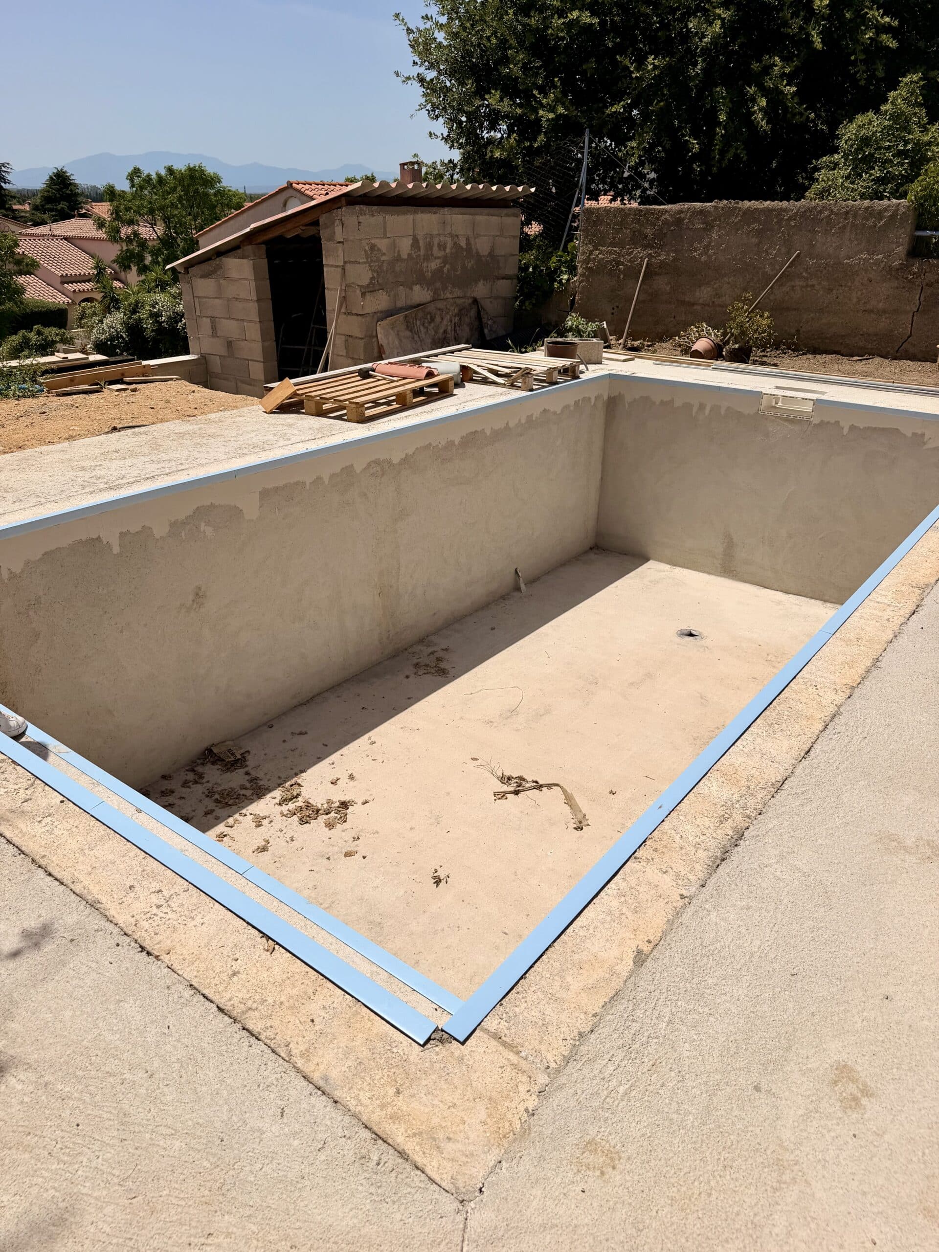 Piscine rectangulaire vide en construction avec bordure bleue et abri de jardin en arrière-plan.