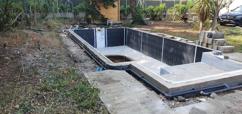 Chantier de piscine creusée avec parois noires et tuyaux PVC bleus visibles dans un jardin en travaux.