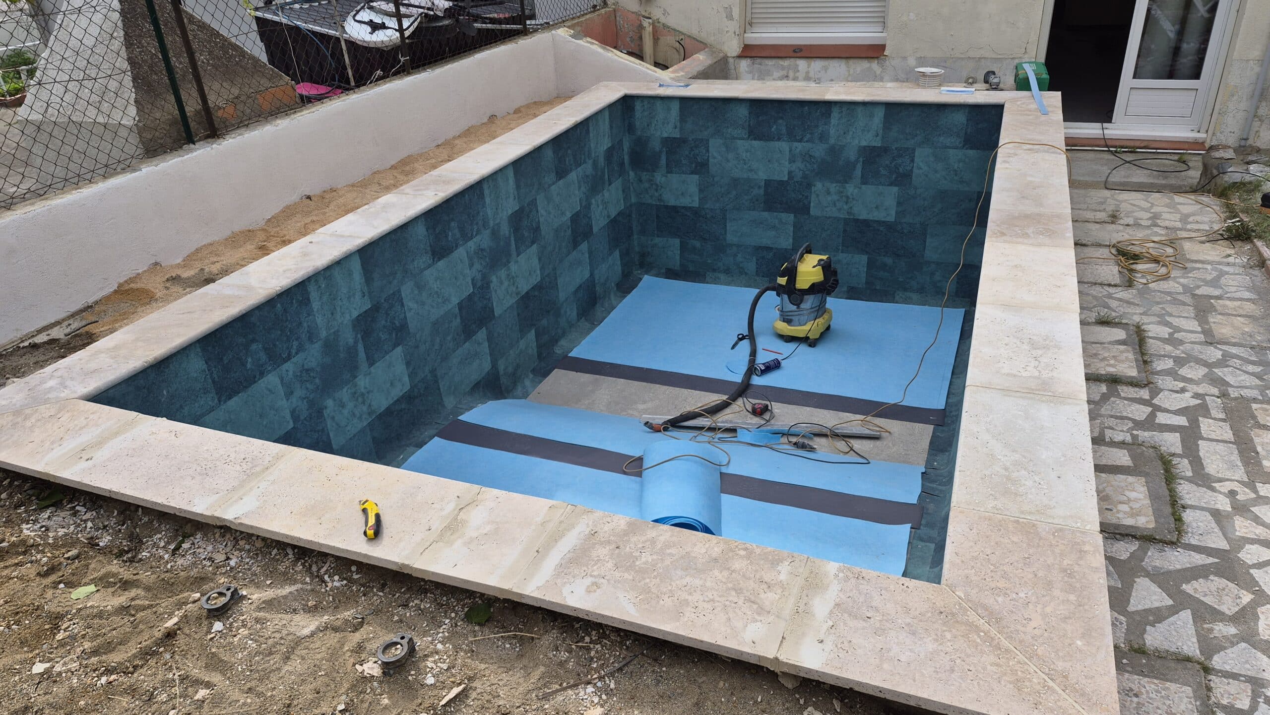 Construction d'une piscine avec liner bleu et margelles en pierre naturelle, aspirateur industriel au fond.