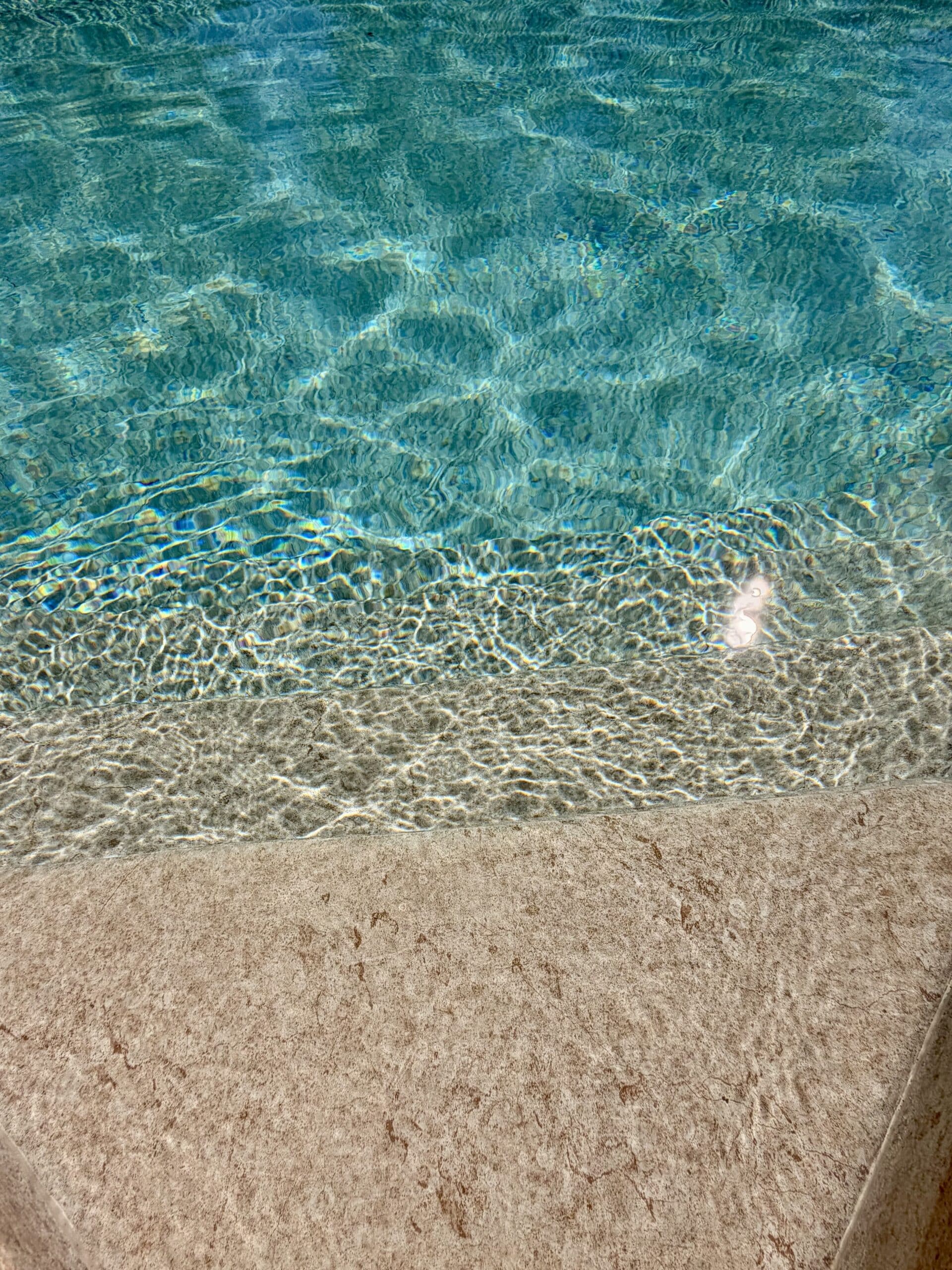 Eau de piscine turquoise scintillante avec reflets du soleil sur des marches immergées en pierre beige.