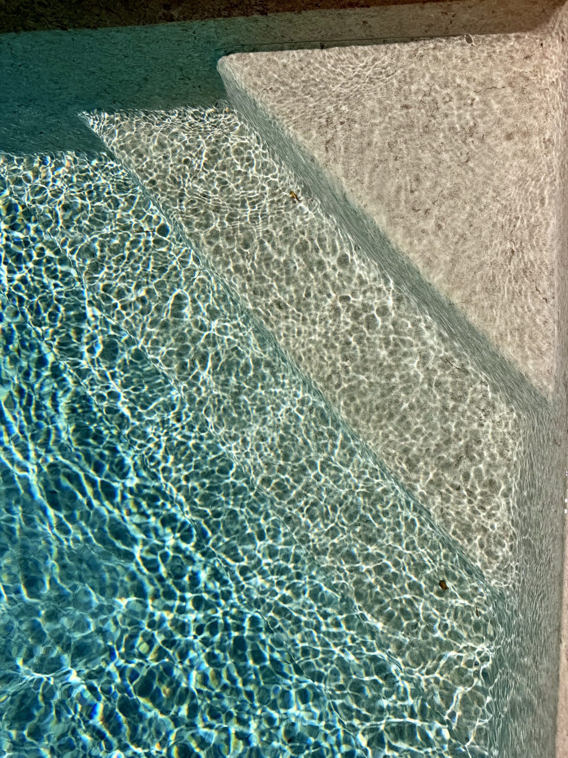Marches de piscine immergées dans l'eau bleue avec reflets de lumière caustiques et textures de pierre claire.