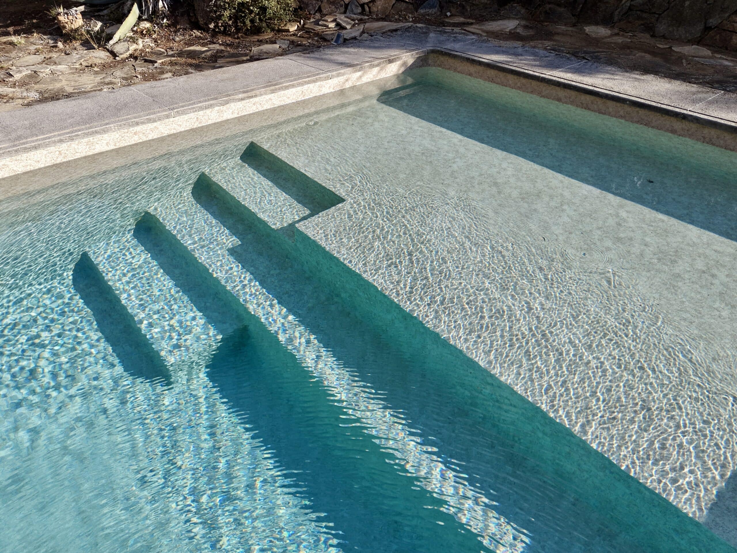 Escalier de piscine immergé dans une eau turquoise claire avec jeux d'ombres et reflets solaires.