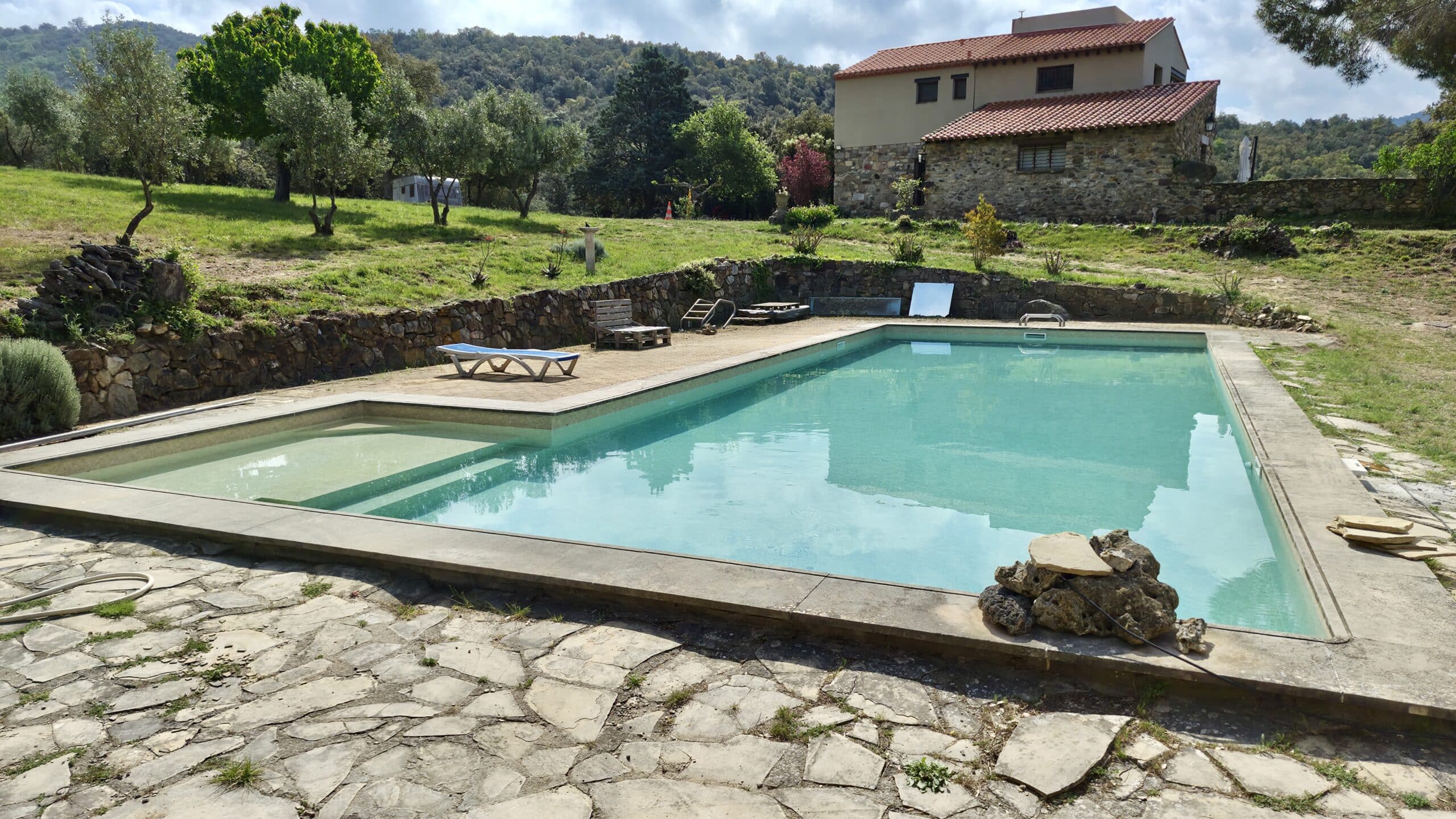 Piscine turquoise et maison de campagne en pierre au pied de collines boisées dans un jardin méditerranéen.