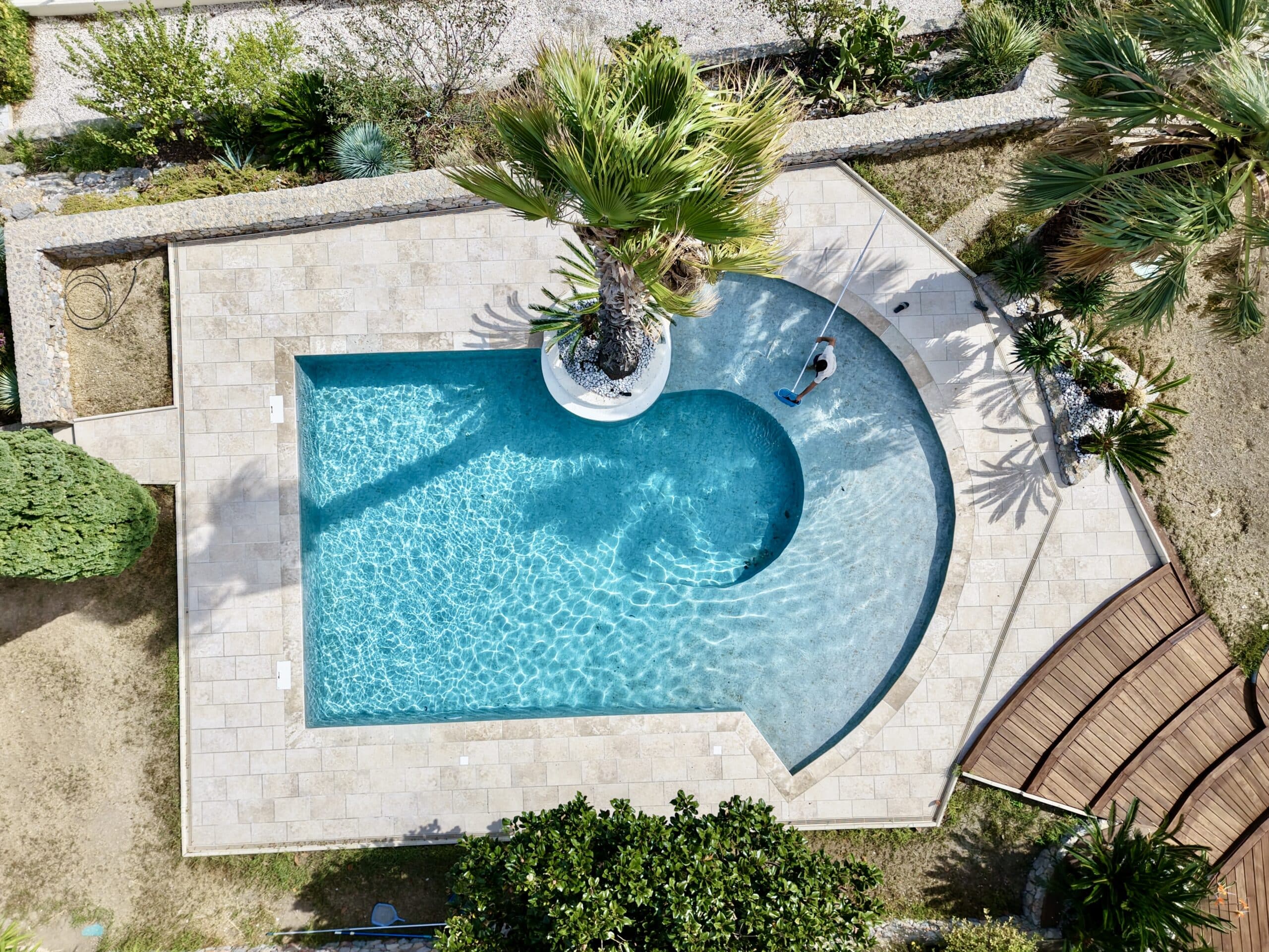 Vue aérienne d'une piscine design avec palmier central en cours de nettoyage dans un jardin méditerranéen.