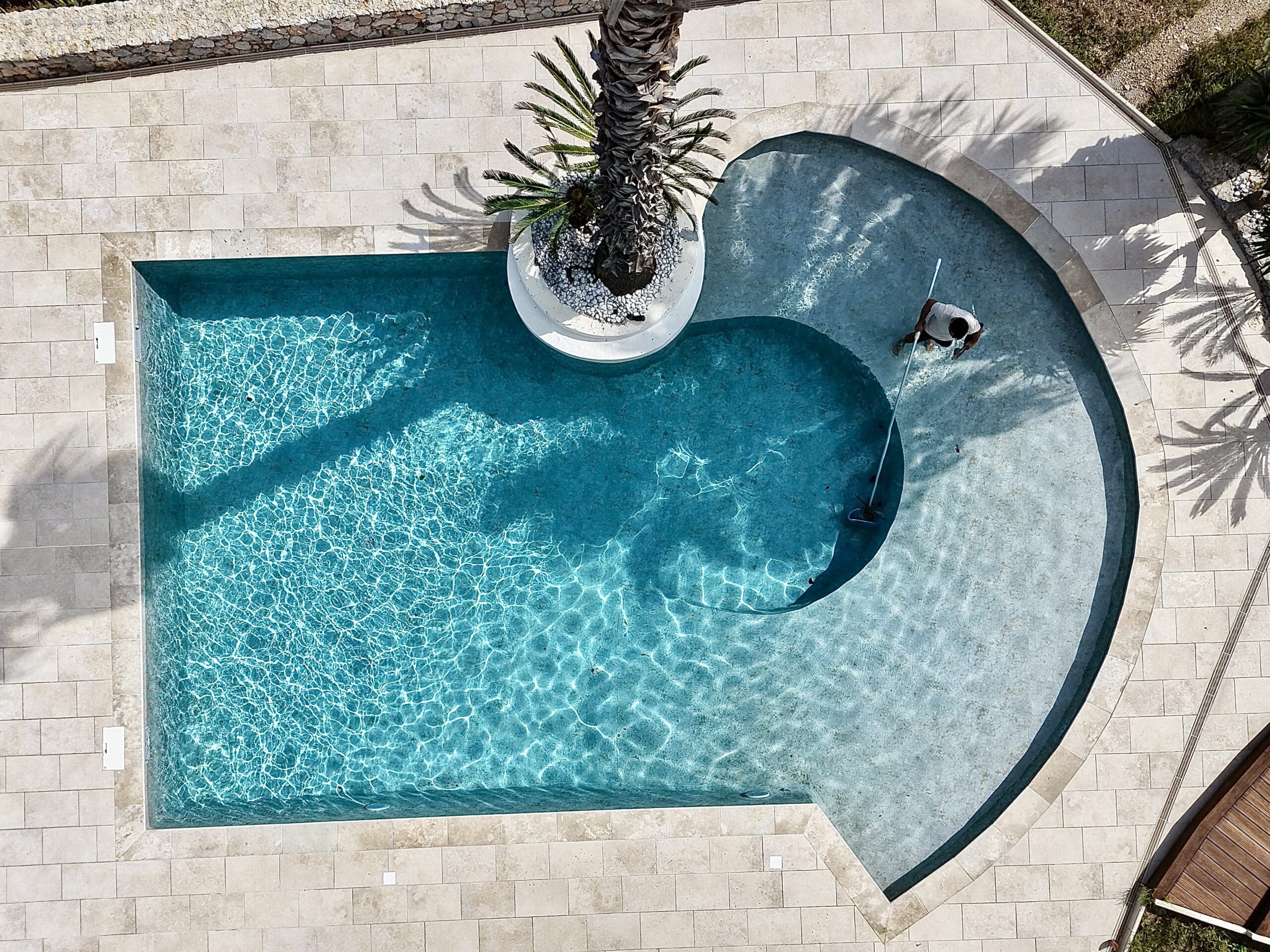 Vue aérienne d'une piscine design bleu clair avec un palmier central et une personne passant l'épuisette.