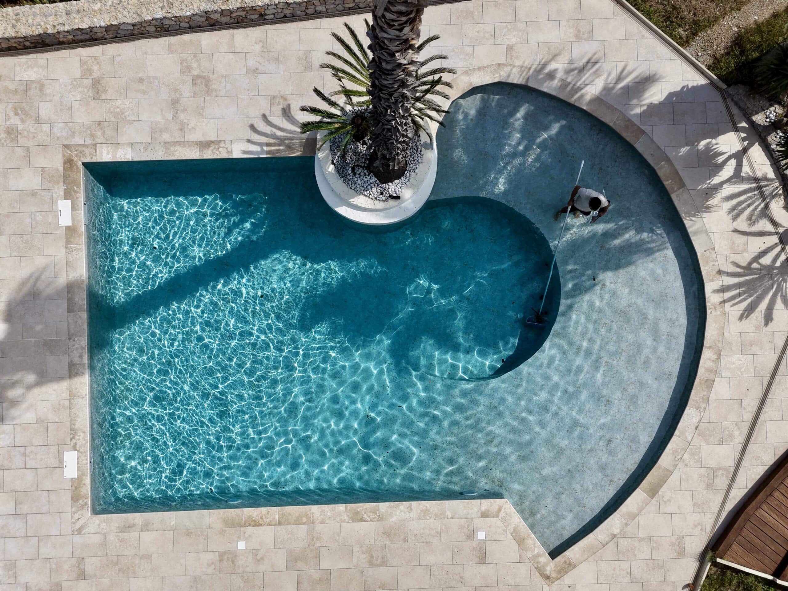 Vue aérienne d'un technicien nettoyant une piscine design avec palmier central et dallage clair au soleil.