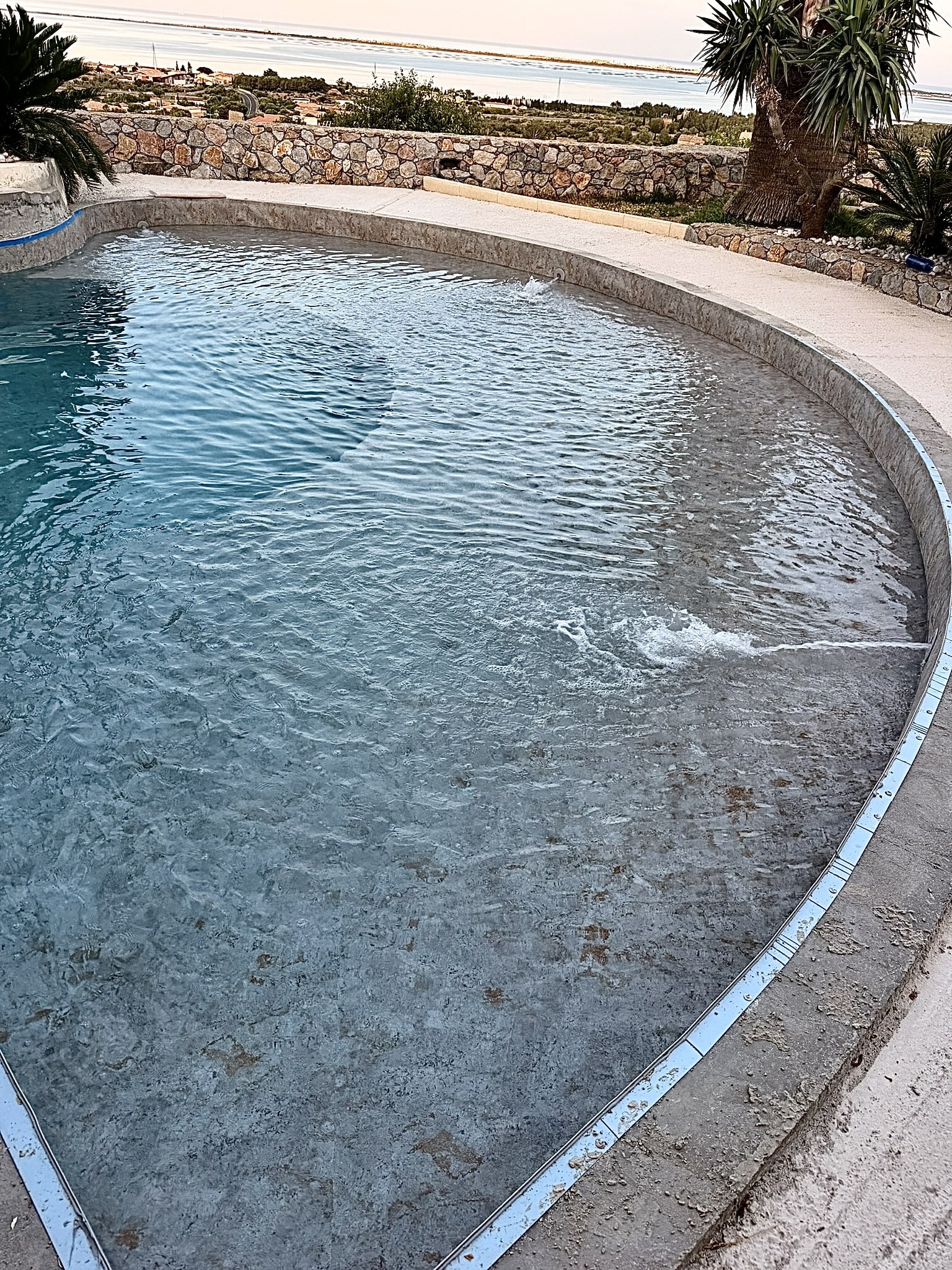 Piscine circulaire à débordement avec eau cristalline, vue panoramique sur la mer et muret en pierre naturelle.