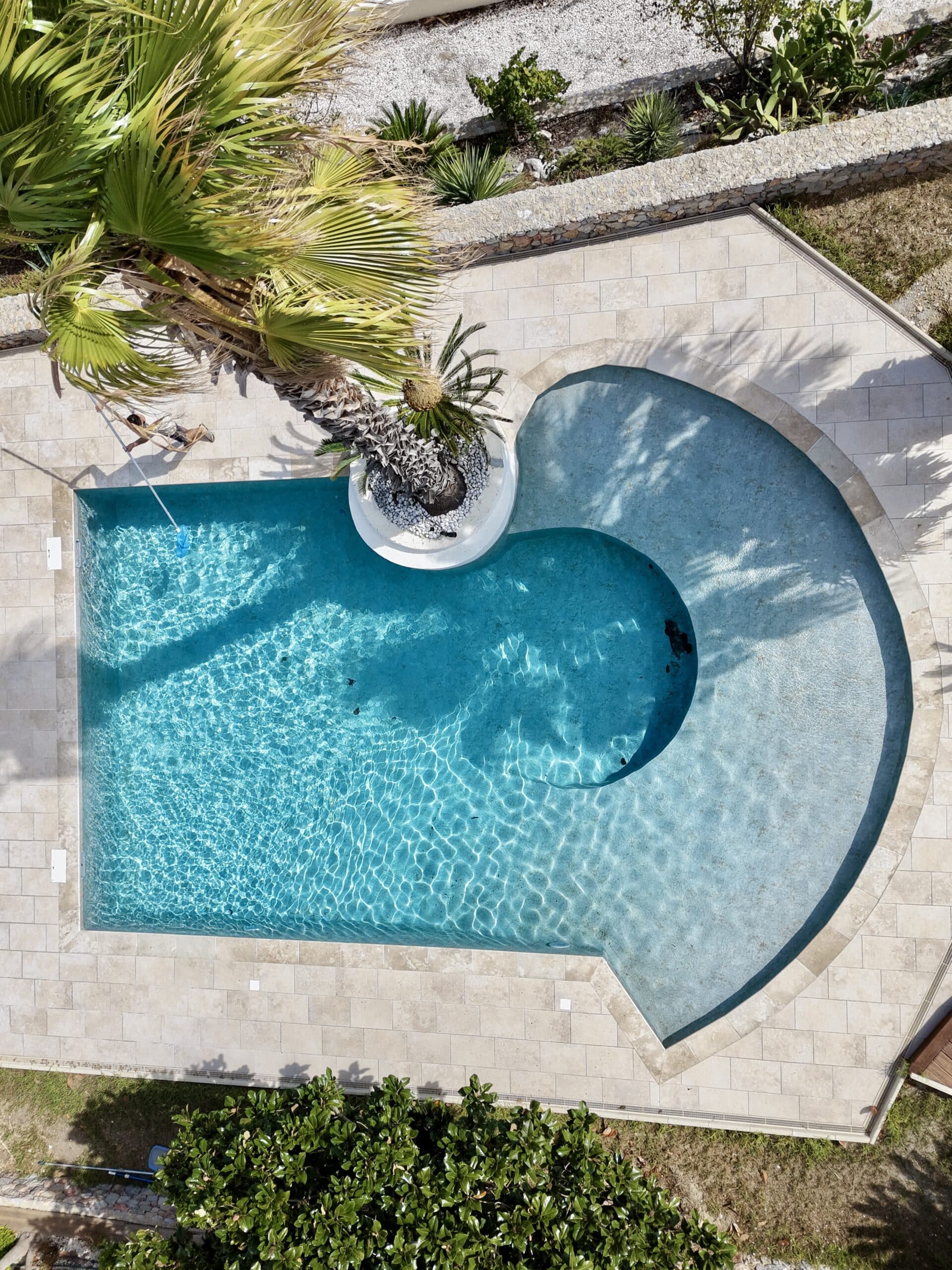Vue aérienne d'une piscine au design original avec palmier intégré, eau cristalline et terrasse en pierre naturelle.