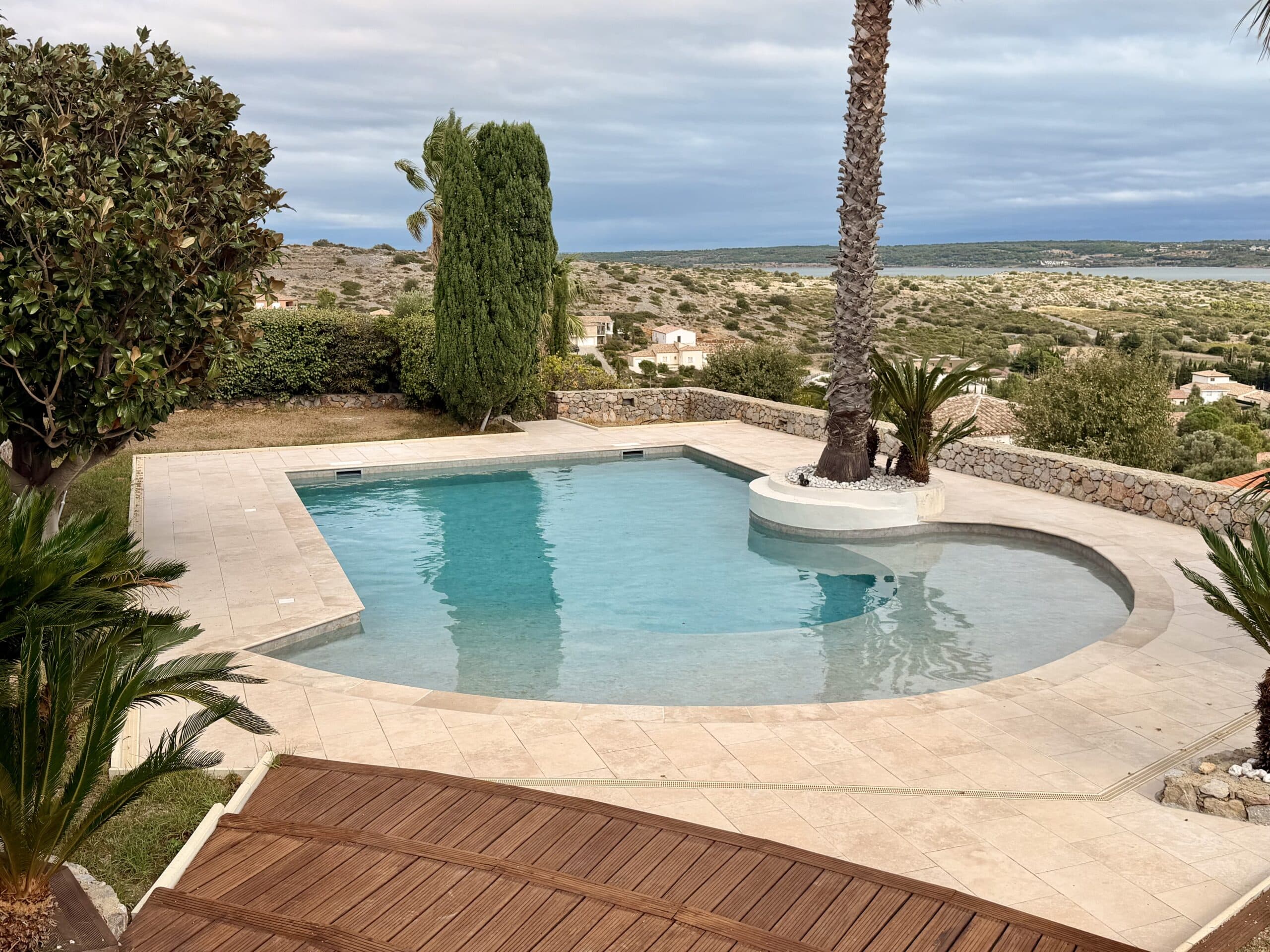 Piscine design avec terrasse en travertin, palmier intégré et vue panoramique sur un paysage méditerranéen et la mer.
