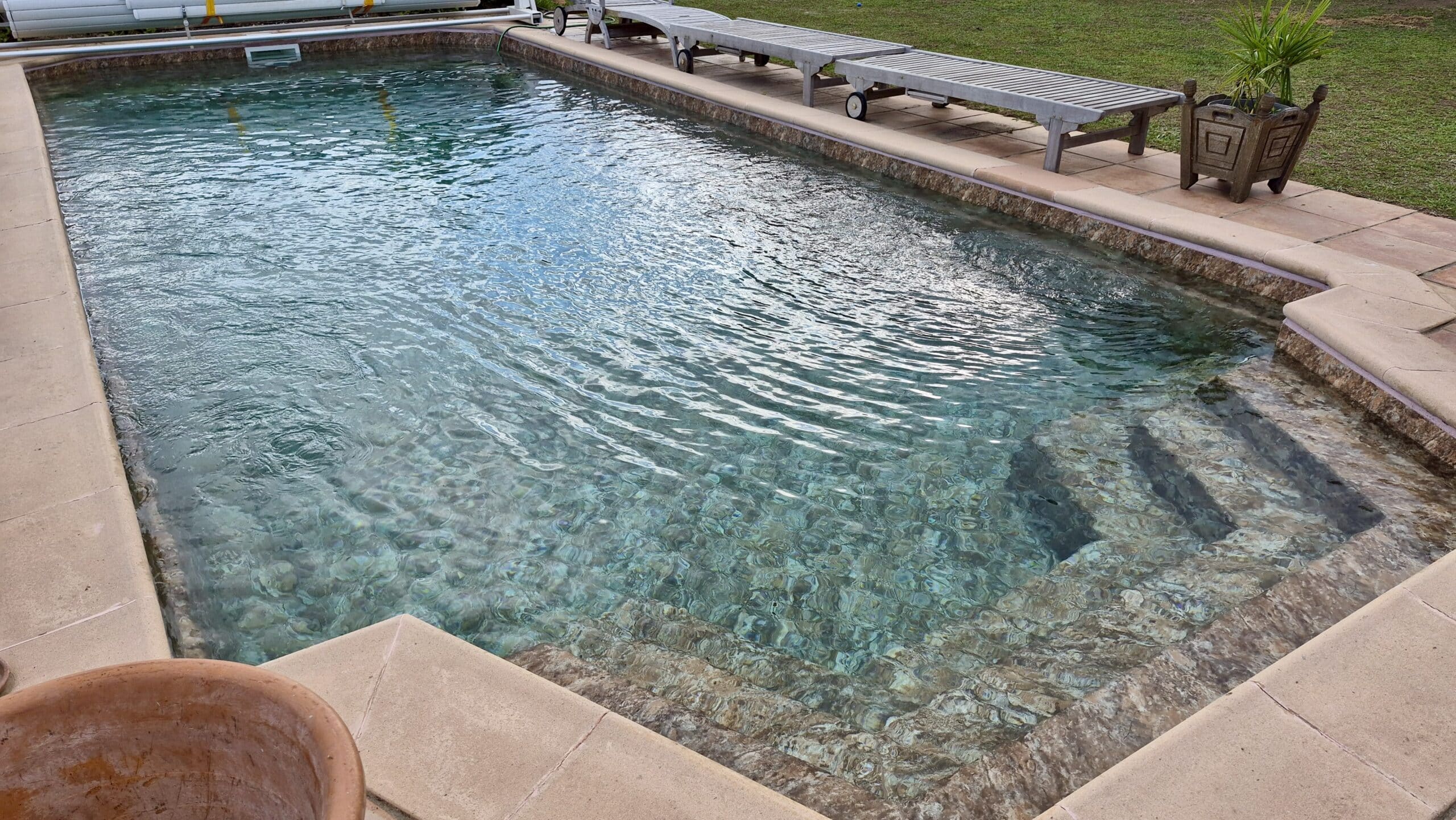 Piscine extérieure à l'eau cristalline avec escalier en pierre et chaises longues sur une terrasse ensoleillée.