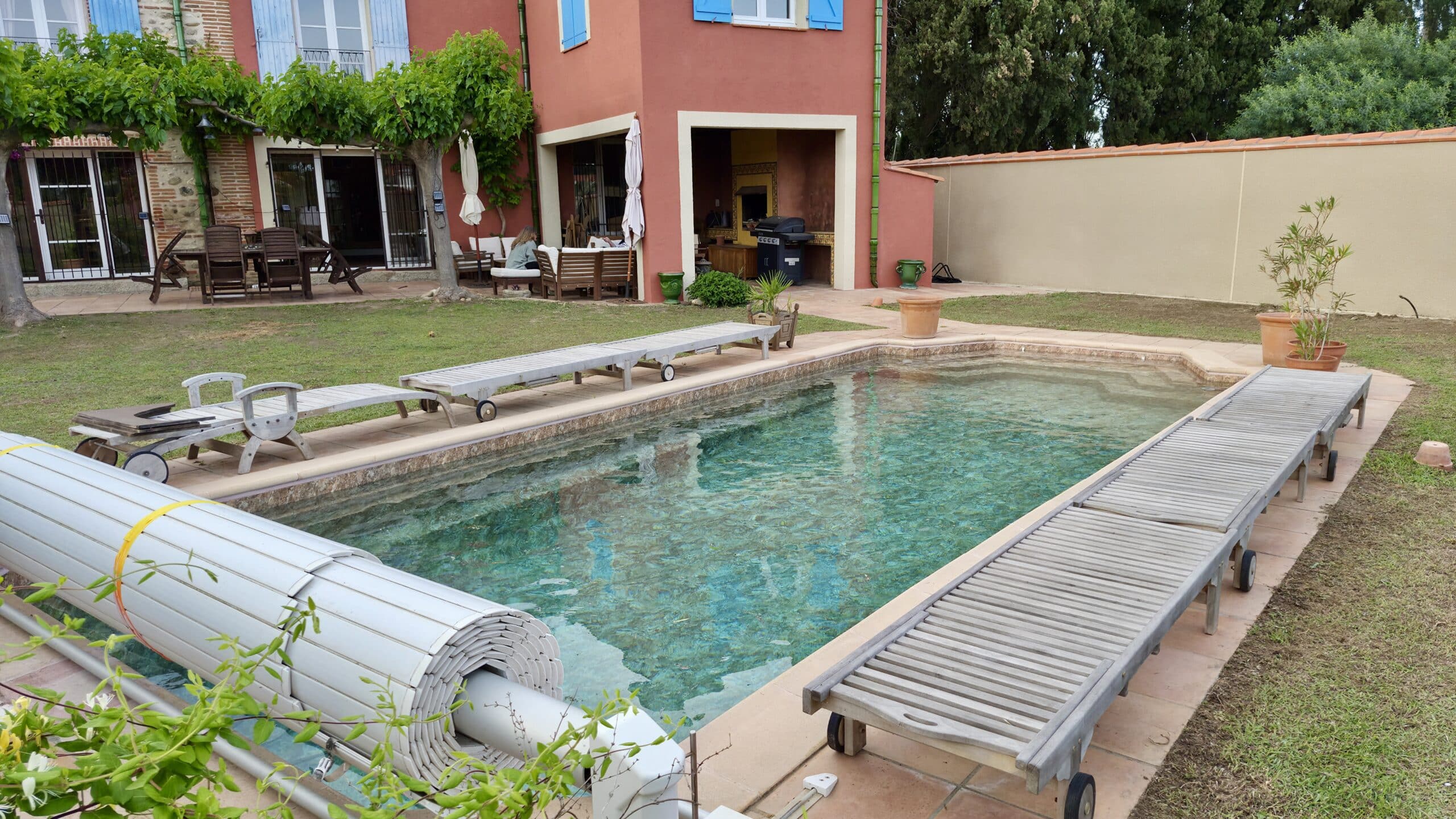 Piscine avec transats en bois devant une maison méditerranéenne en terre cuite avec terrasse et jardin arboré.