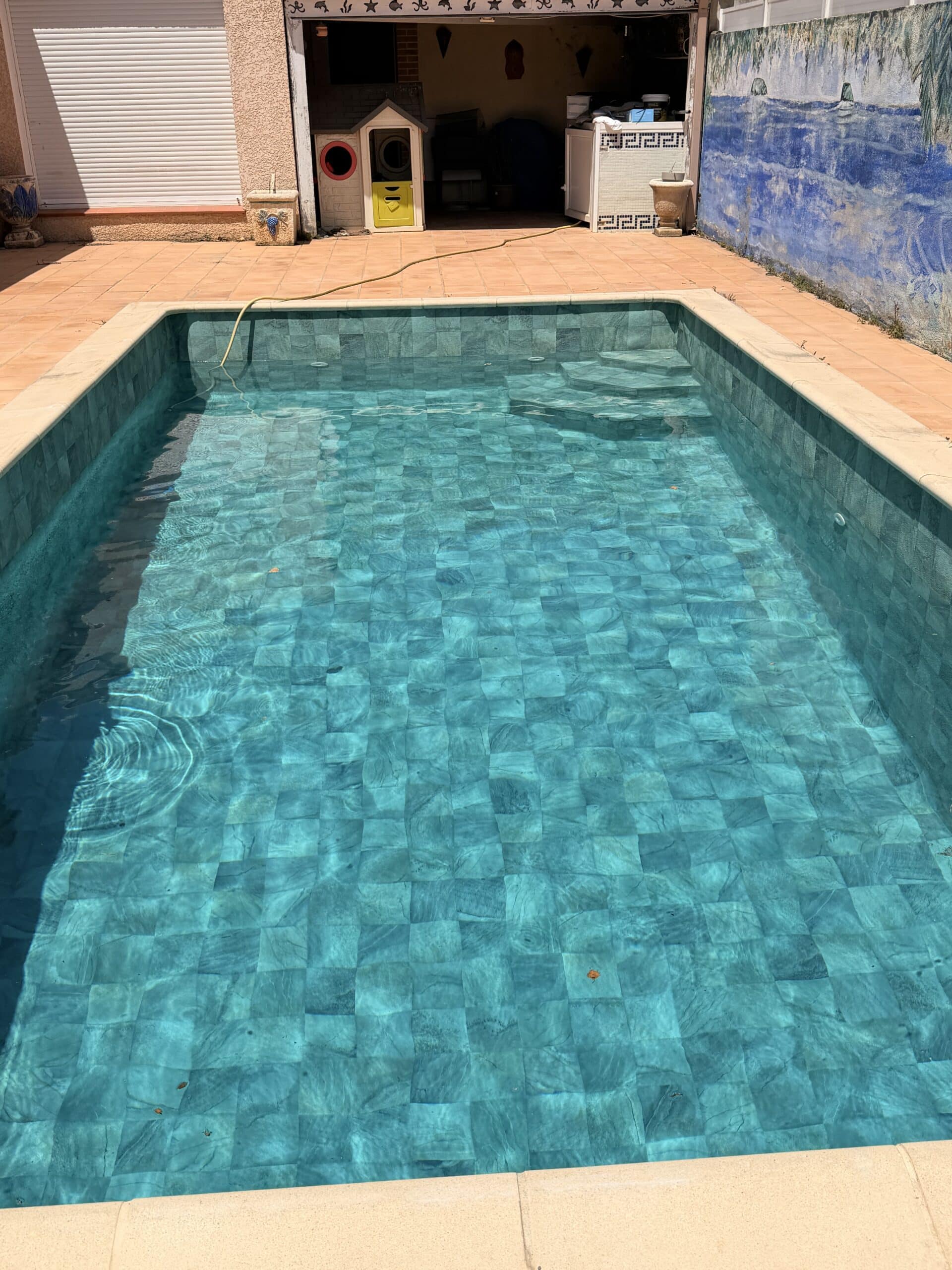 Piscine rectangulaire aux eaux turquoises, carrelage en mosaïque, terrasse ensoleillée et aire de jeux au fond.