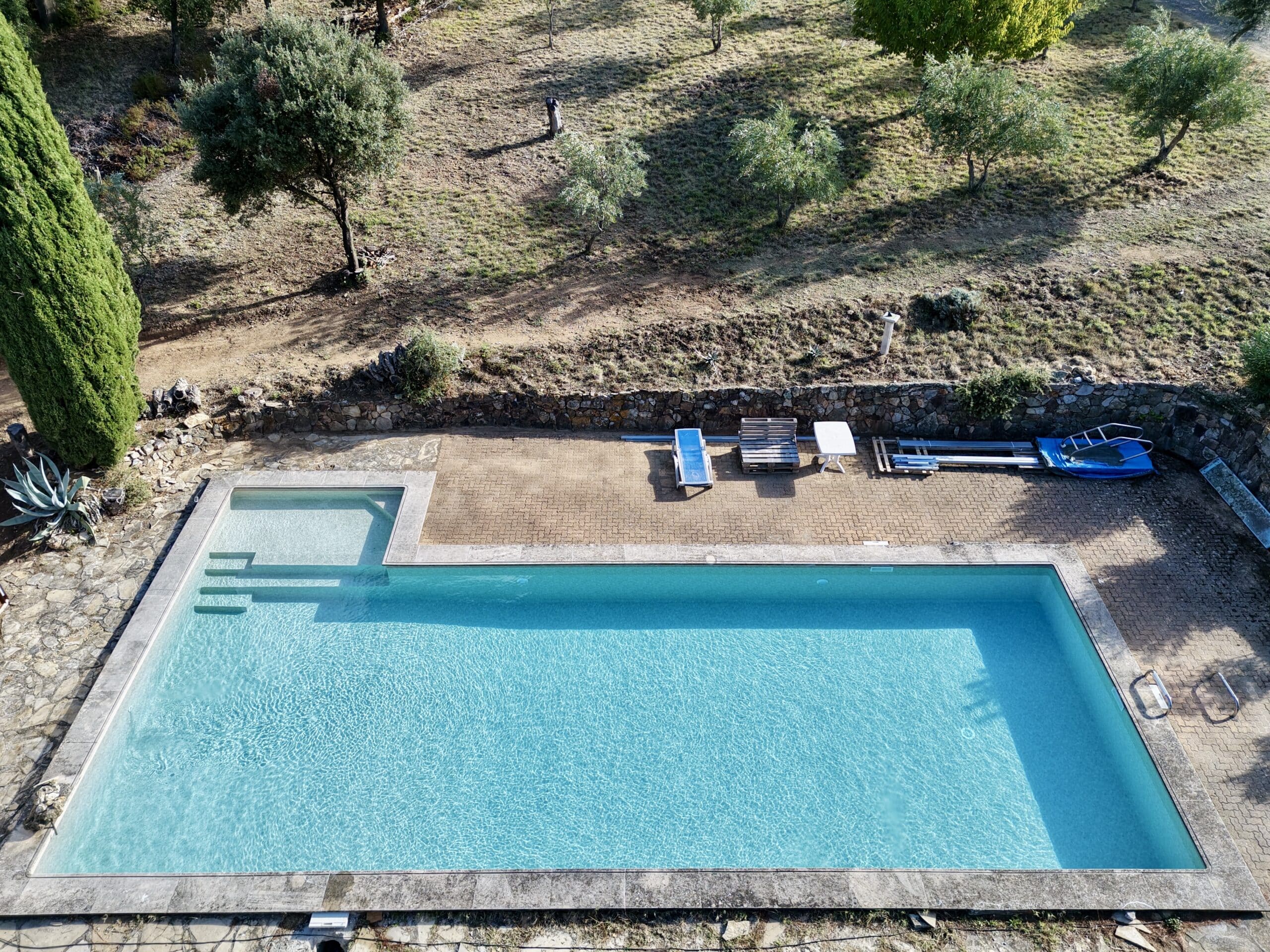 Vue aérienne d'une grande piscine rectangulaire entourée de dalles en pierre et d'un jardin avec des oliviers.