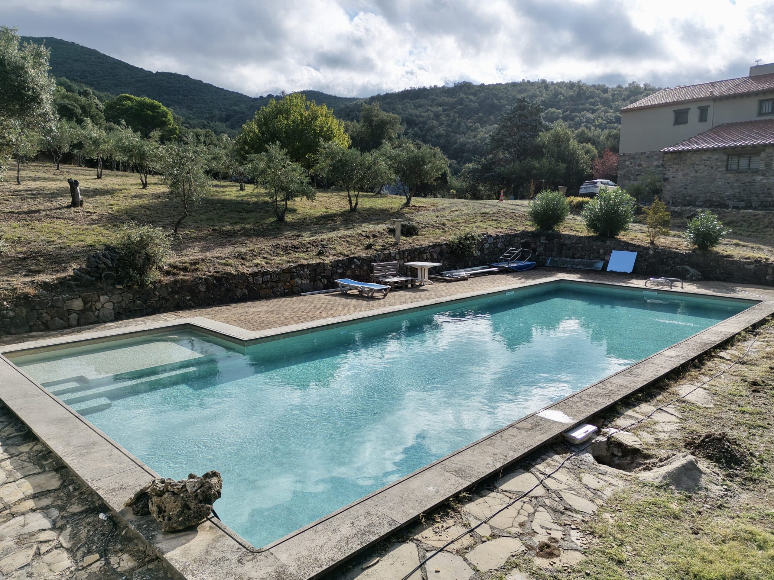 Piscine extérieure avec vue sur les montagnes, terrasse en pierre et oliviers sous un ciel nuageux en campagne.