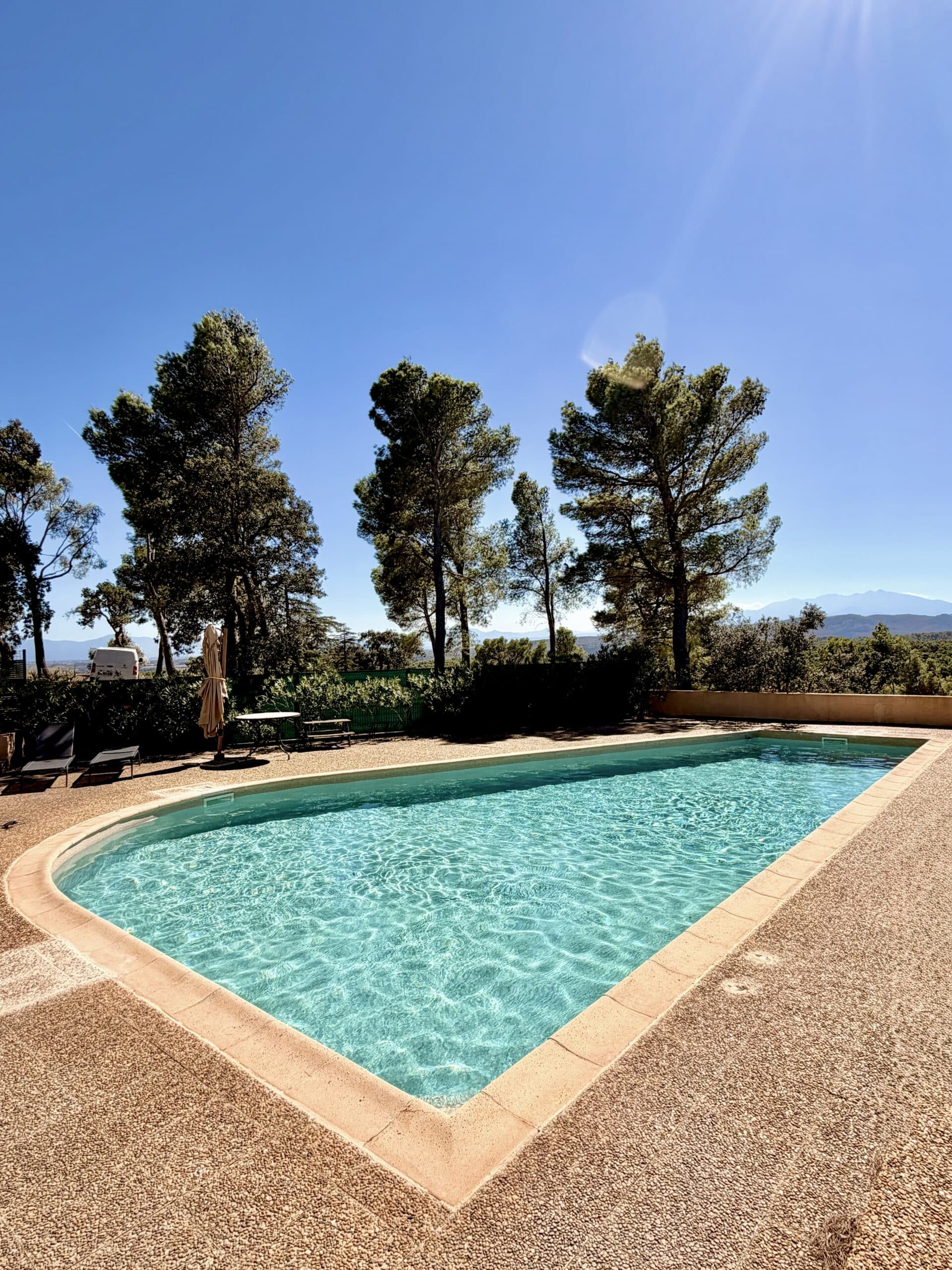 Piscine turquoise entourée de pins sous un ciel bleu avec vue sur les montagnes par une journée ensoleillée.