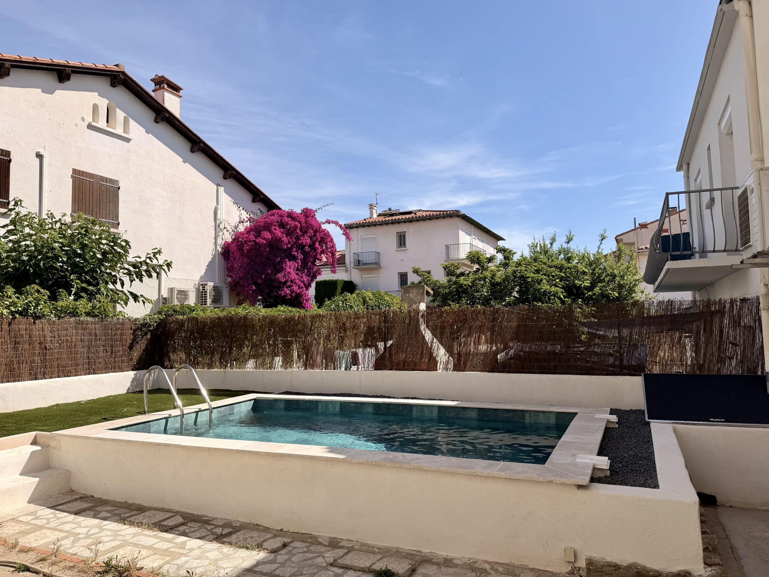 Petite piscine moderne dans un jardin ensoleillé avec bougainvillier rose et maisons blanches méditerranéennes.