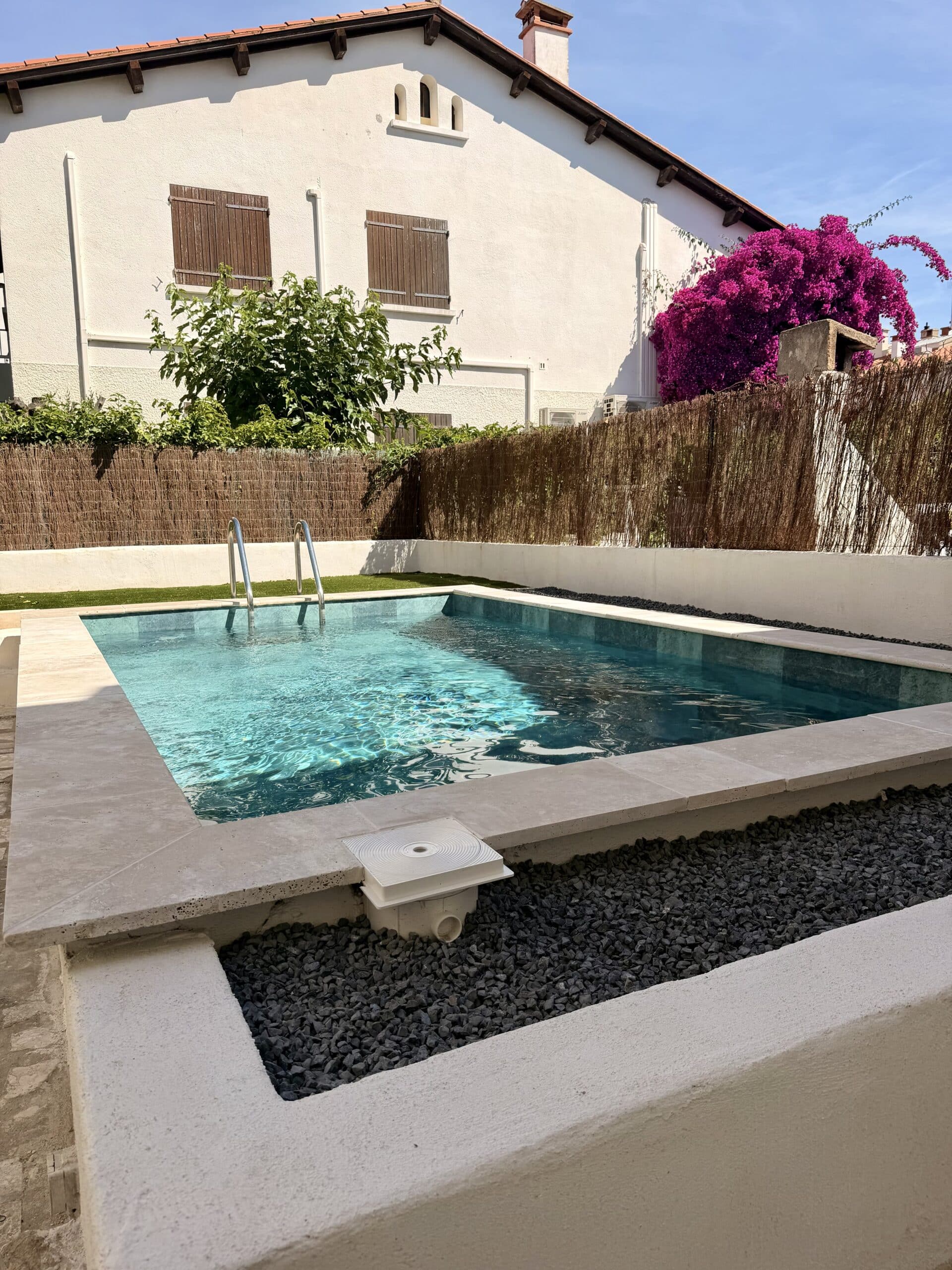 Petite piscine moderne avec échelle, clôture en brande et bougainvillier rose devant une maison blanche.