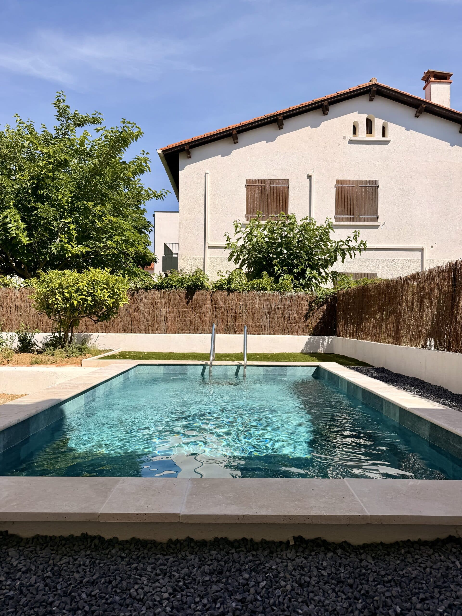 Piscine rectangulaire moderne dans un jardin ensoleillé avec maison blanche et ciel bleu.