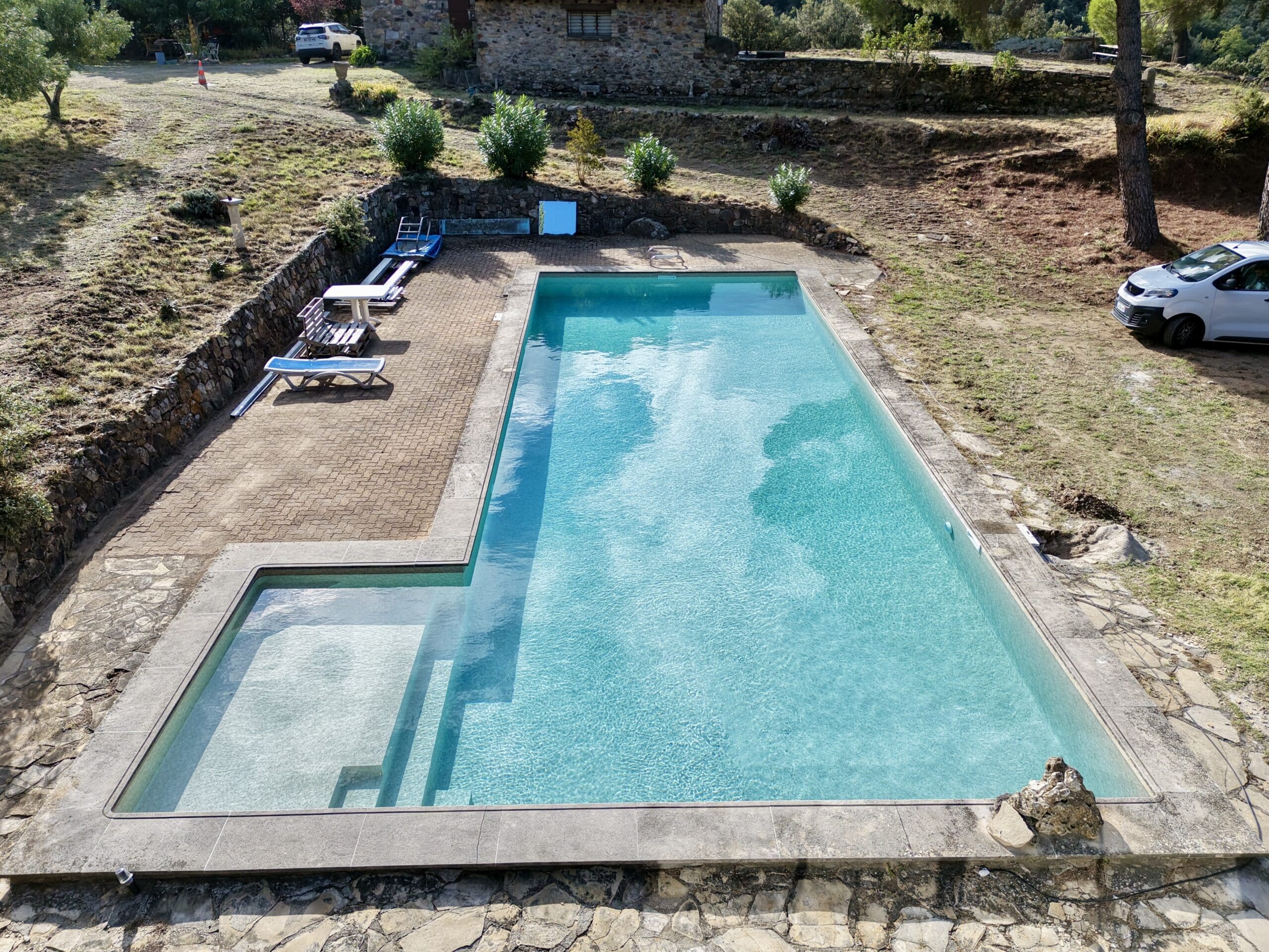 Piscine rectangulaire turquoise avec escaliers intégrés dans un jardin rustique avec terrasse en pierre et transats.