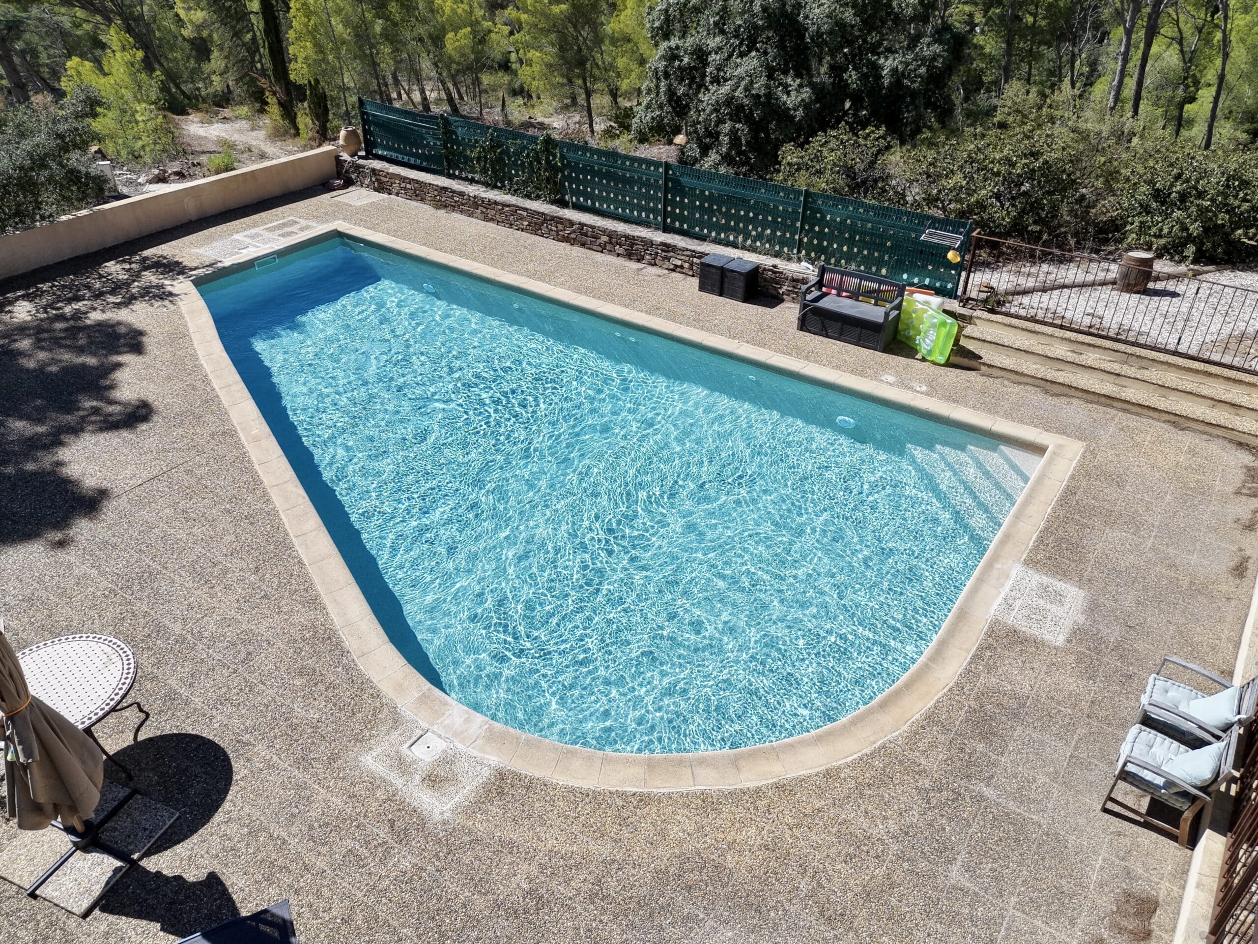 Piscine aux eaux turquoises entourée d'une terrasse en pierre et de verdure sous le soleil.
