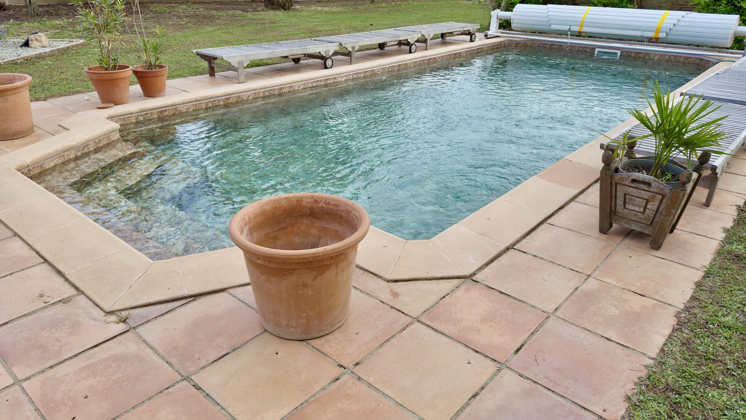 Piscine de jardin entourée d'une terrasse en dalles, pots en terre cuite et chaises longues en bois.