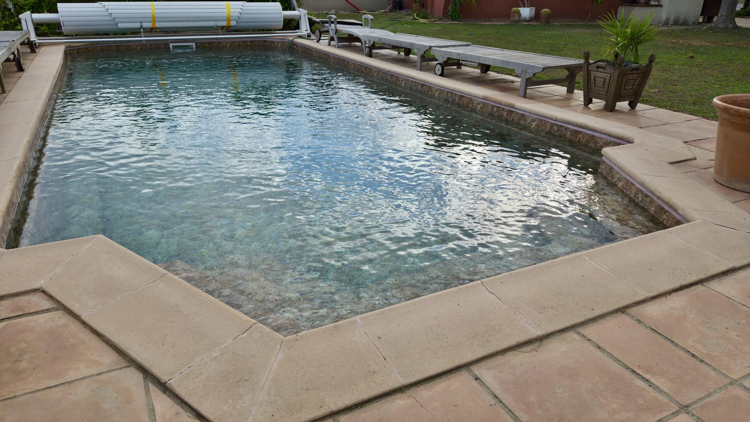 Piscine aux eaux claires avec margelles en pierre, transats en bois et volet roulant.