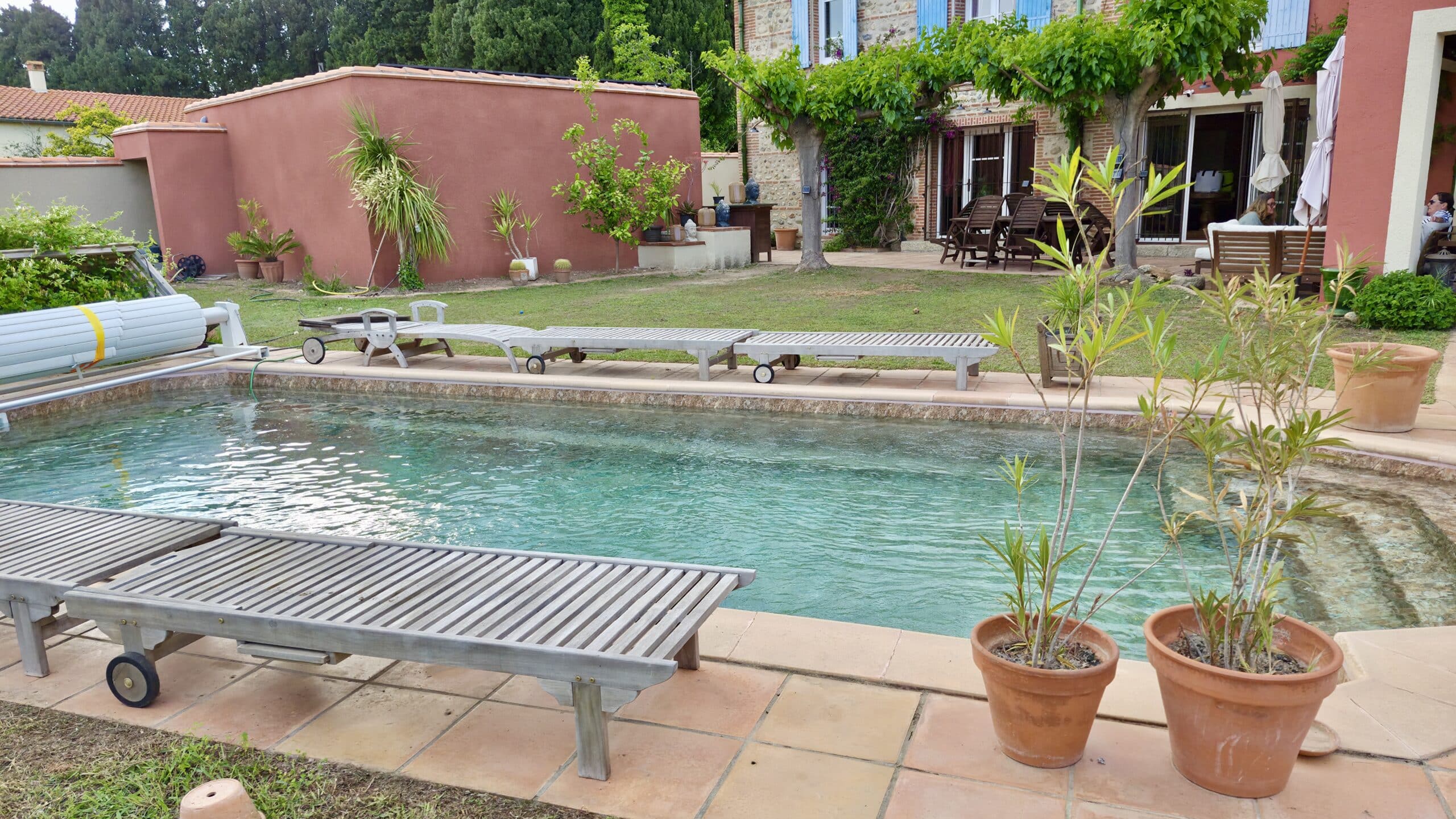 Piscine extérieure avec transats en bois, pots en terre cuite et maison méditerranéenne en arrière-plan.