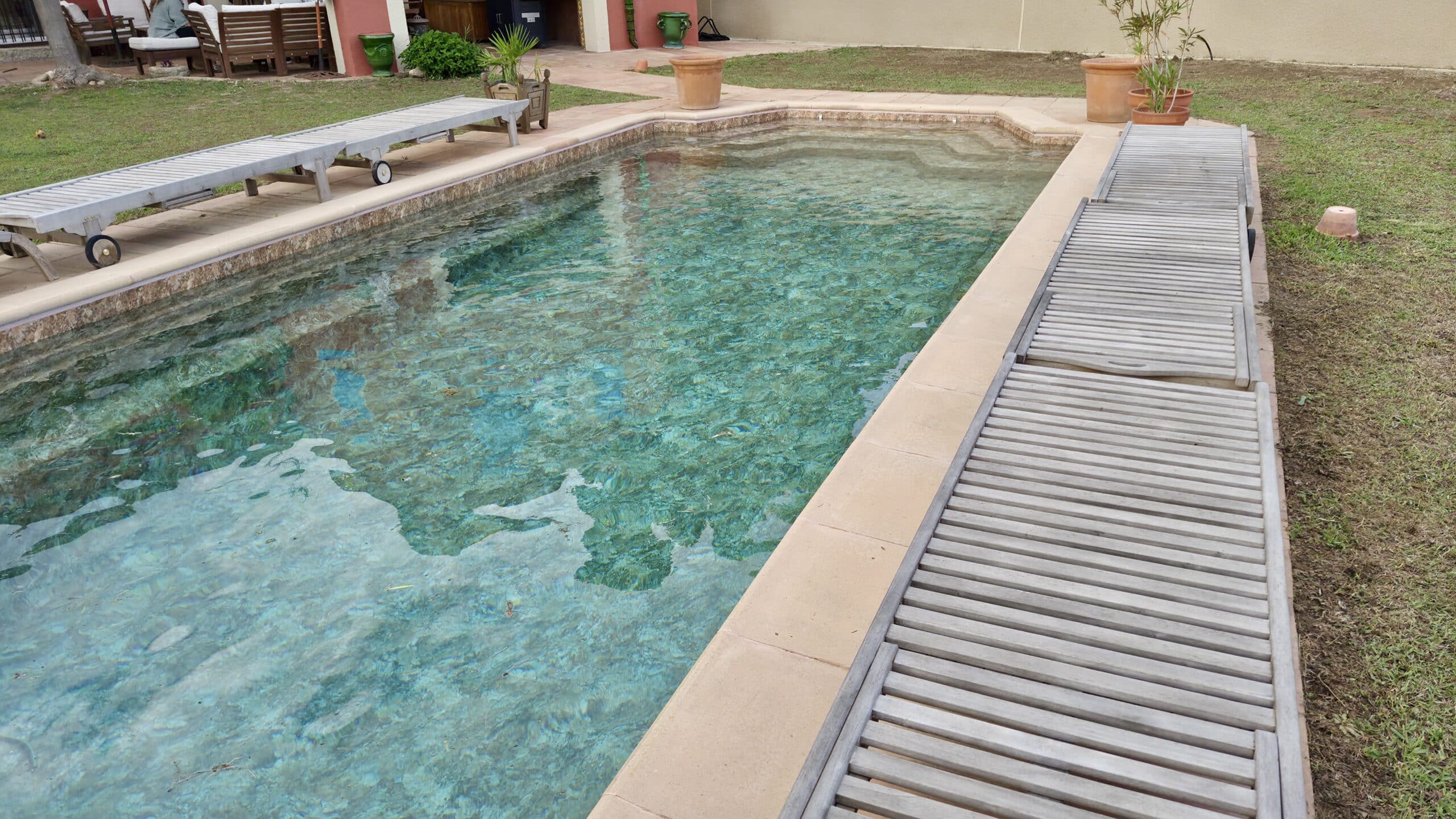 Piscine turquoise bordée de transats en bois gris et d'un jardin avec pelouse et pots en terre cuite.