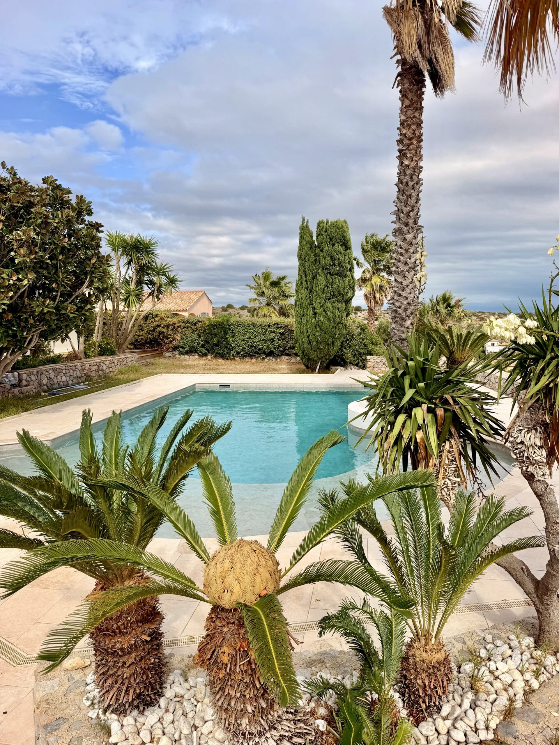 Piscine turquoise dans un jardin tropical avec palmiers et cyprès sous un ciel nuageux.
