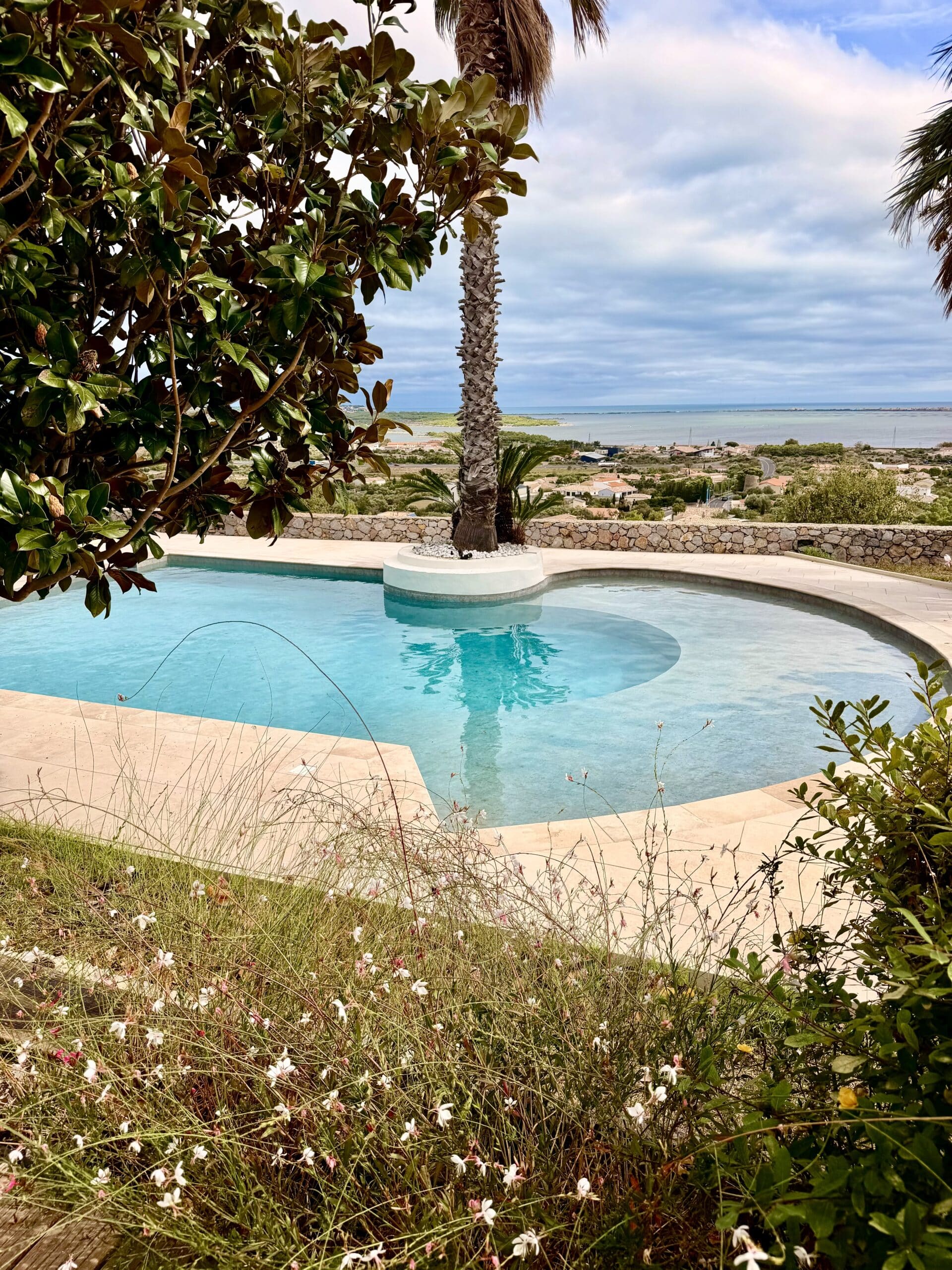 Piscine courbée avec palmier central et vue panoramique sur la mer Méditerranée depuis un jardin fleuri.