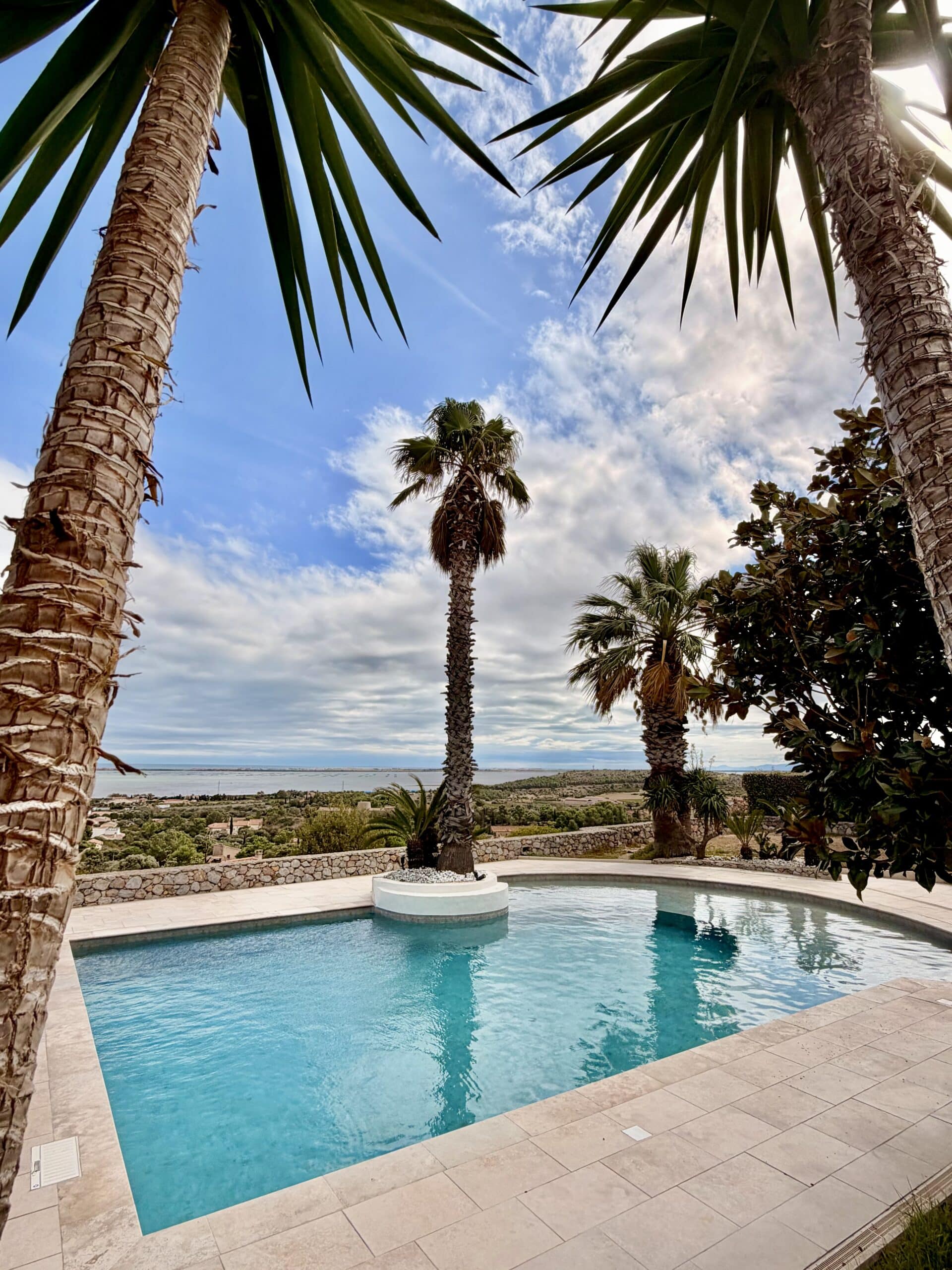 Piscine luxueuse entourée de palmiers avec vue imprenable sur la mer et le paysage méditerranéen sous un ciel bleu.