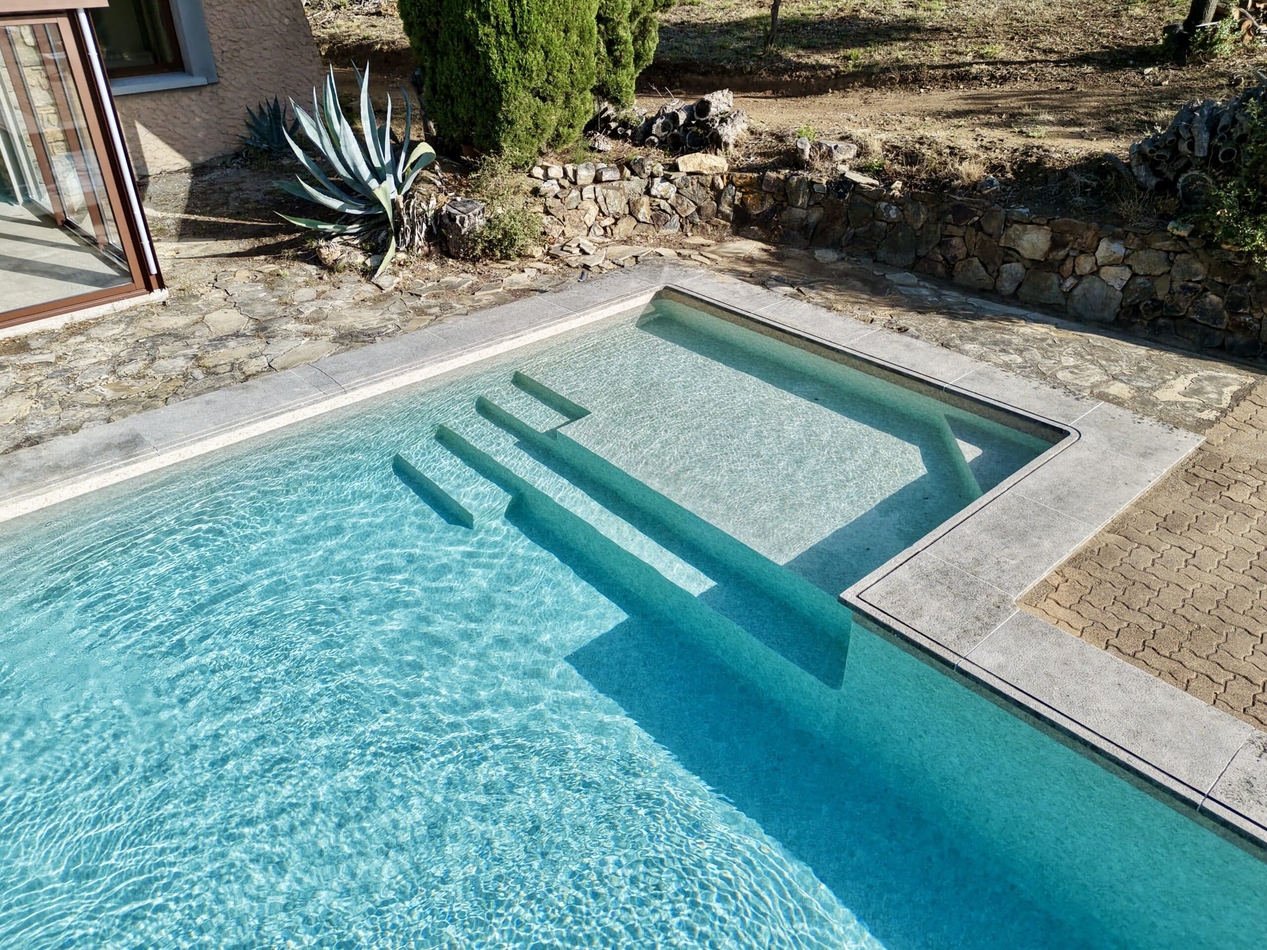 Piscine moderne avec escalier immergé, margelles en pierre grise et eau turquoise dans un jardin ensoleillé.