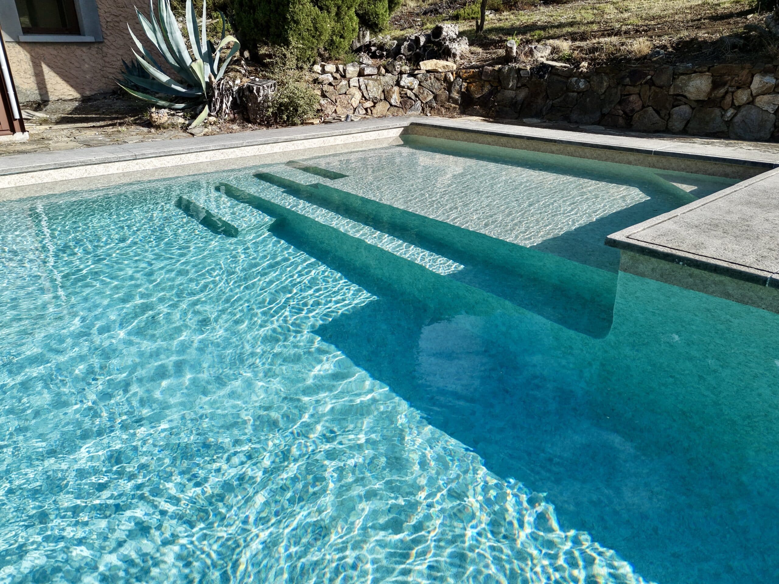 Piscine en pierre naturelle avec escalier immergé, eau cristalline et agave dans un jardin méditerranéen ensoleillé.