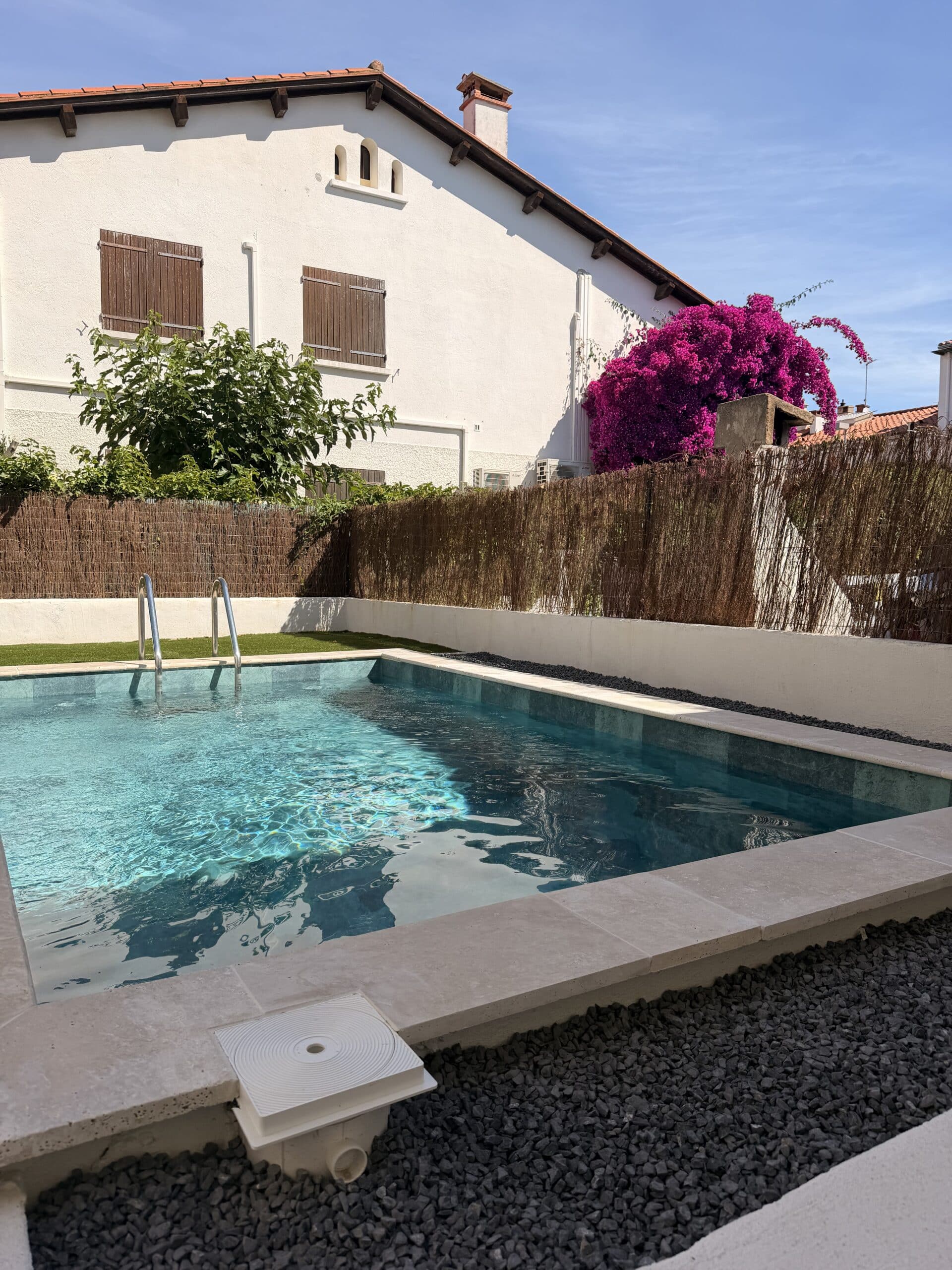 Piscine rectangulaire moderne dans un jardin avec maison blanche, volets marron et bougainvillier rose éclatant.