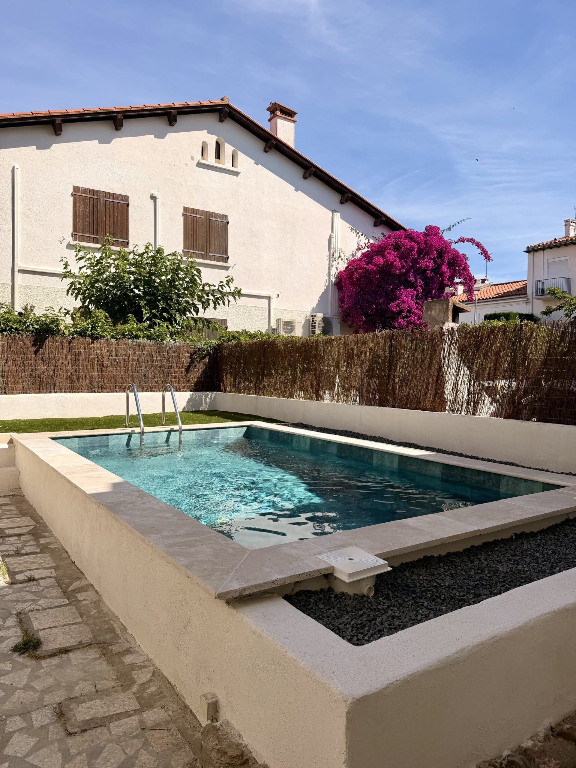 Piscine moderne avec margelles en pierre devant une maison blanche et bougainvillée violette éclatante.