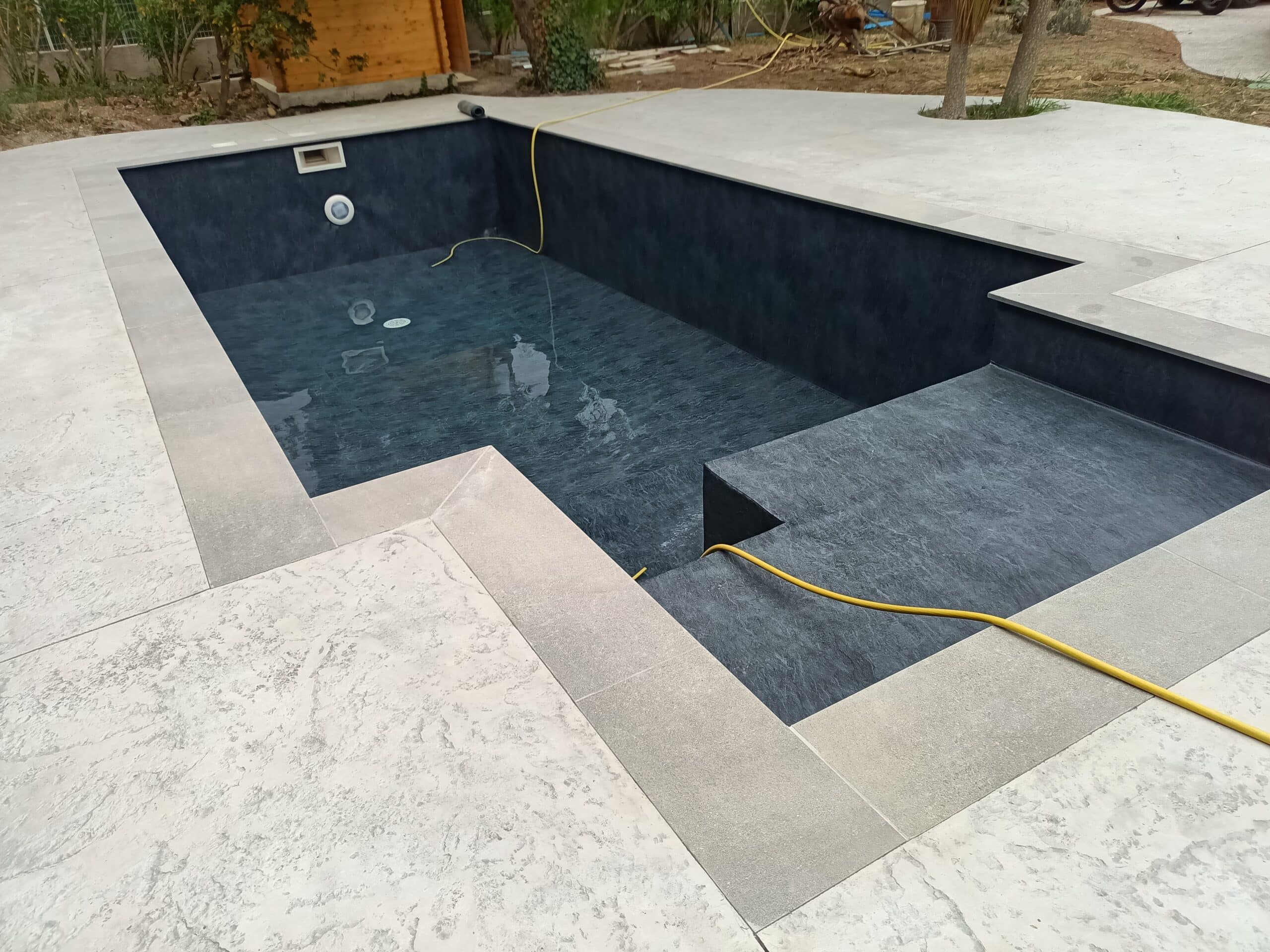 Piscine design au liner gris anthracite avec entourage en béton imprimé beige et tuyau de remplissage jaune.
