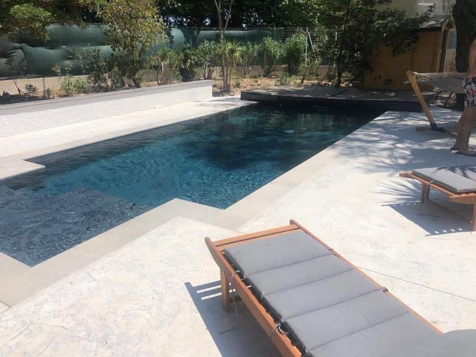 Piscine rectangulaire moderne avec eau bleu foncé, terrasse claire et transats en bois dans un jardin arboré.