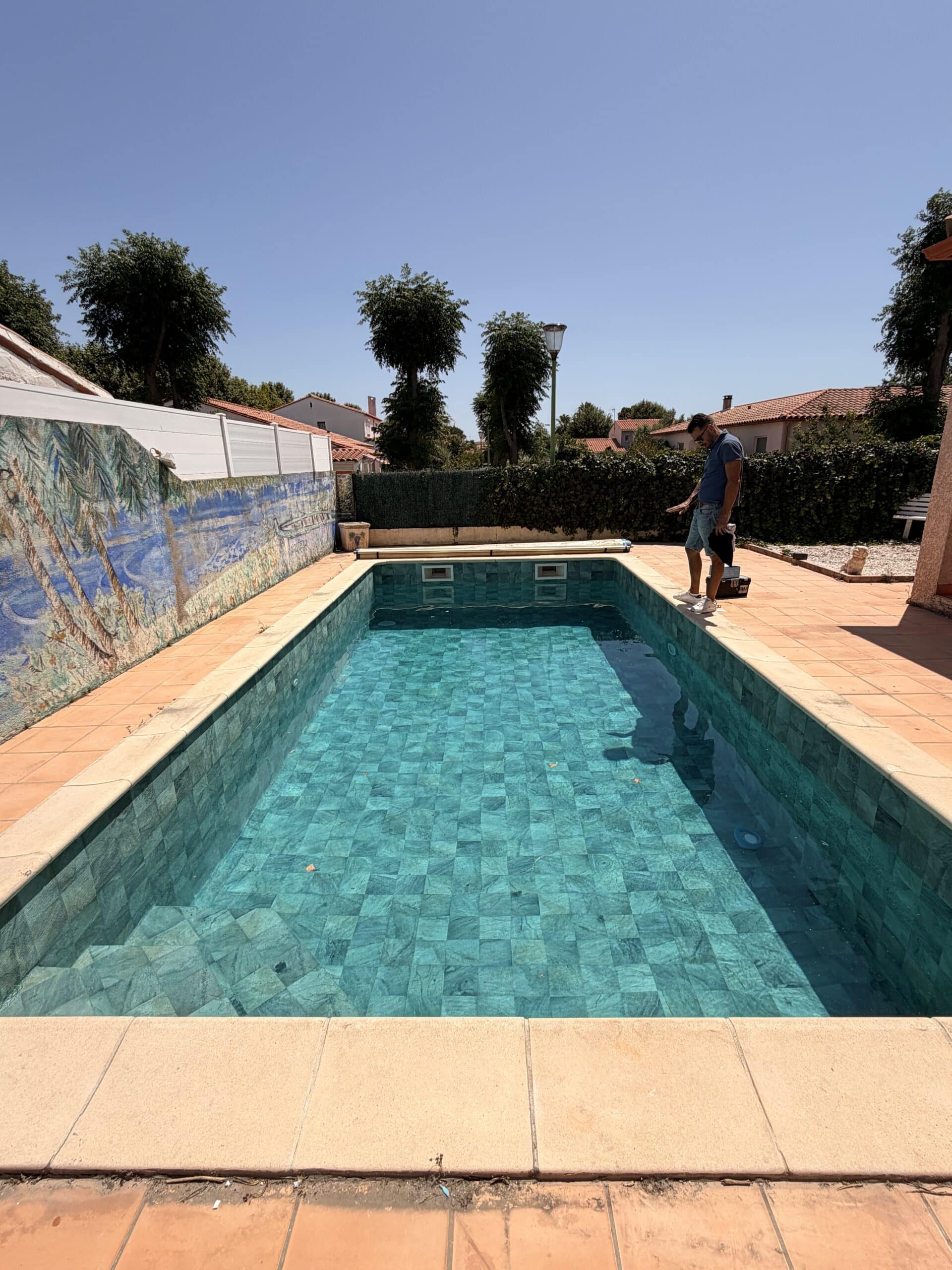 Piscine rectangulaire en mosaïque turquoise avec terrasse en terre cuite et homme debout sur la margelle sous un ciel bleu.