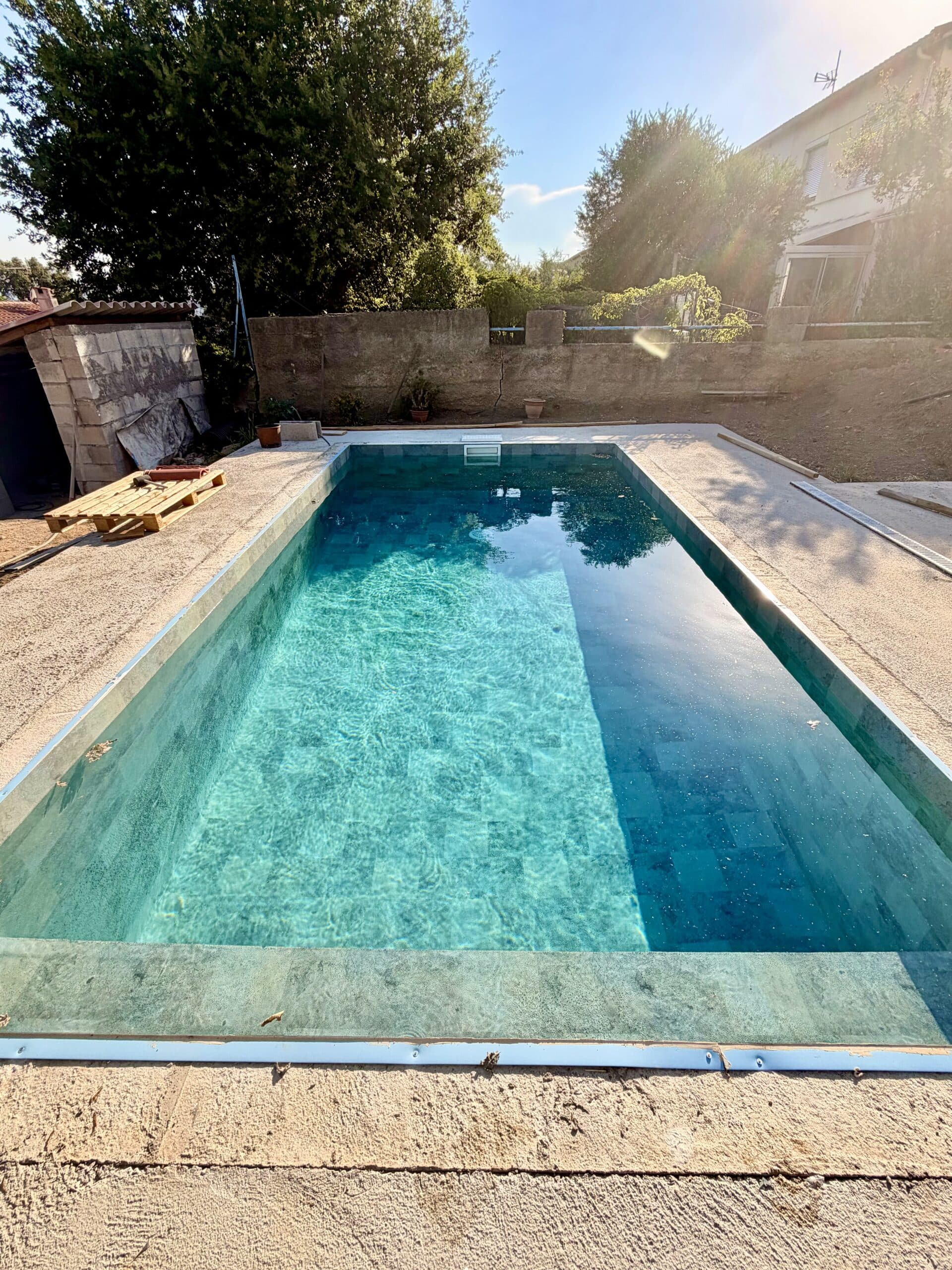 Piscine rectangulaire moderne avec revêtement turquoise et margelles en pierre dans un jardin ensoleillé.