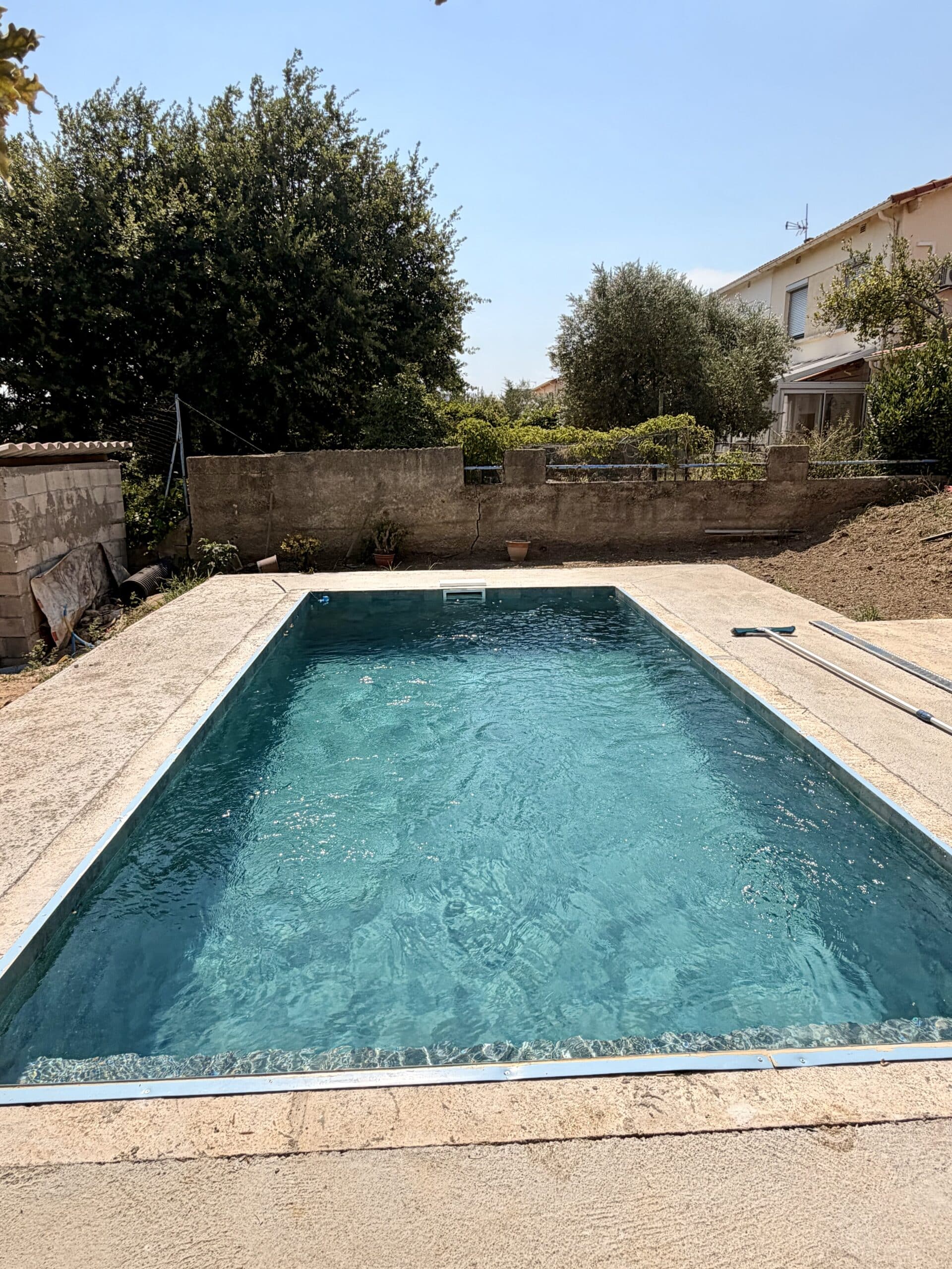 Une piscine rectangulaire à l'eau bleue cristalline entourée d'une terrasse en béton sous un ciel dégagé et ensoleillé.